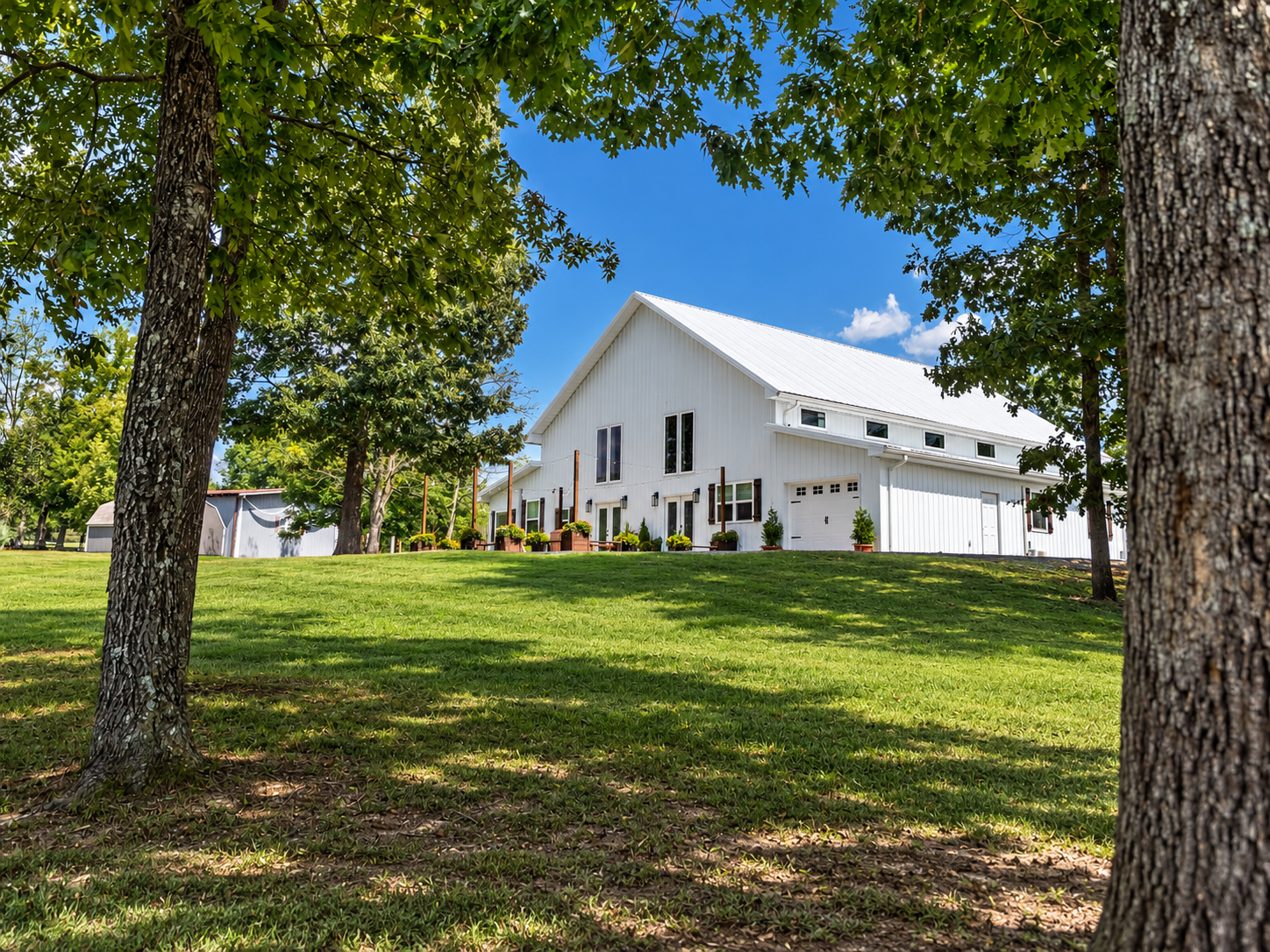 A white barn with a metal roof, surrounded by trees and green grass on a sunny day with blue sky.
