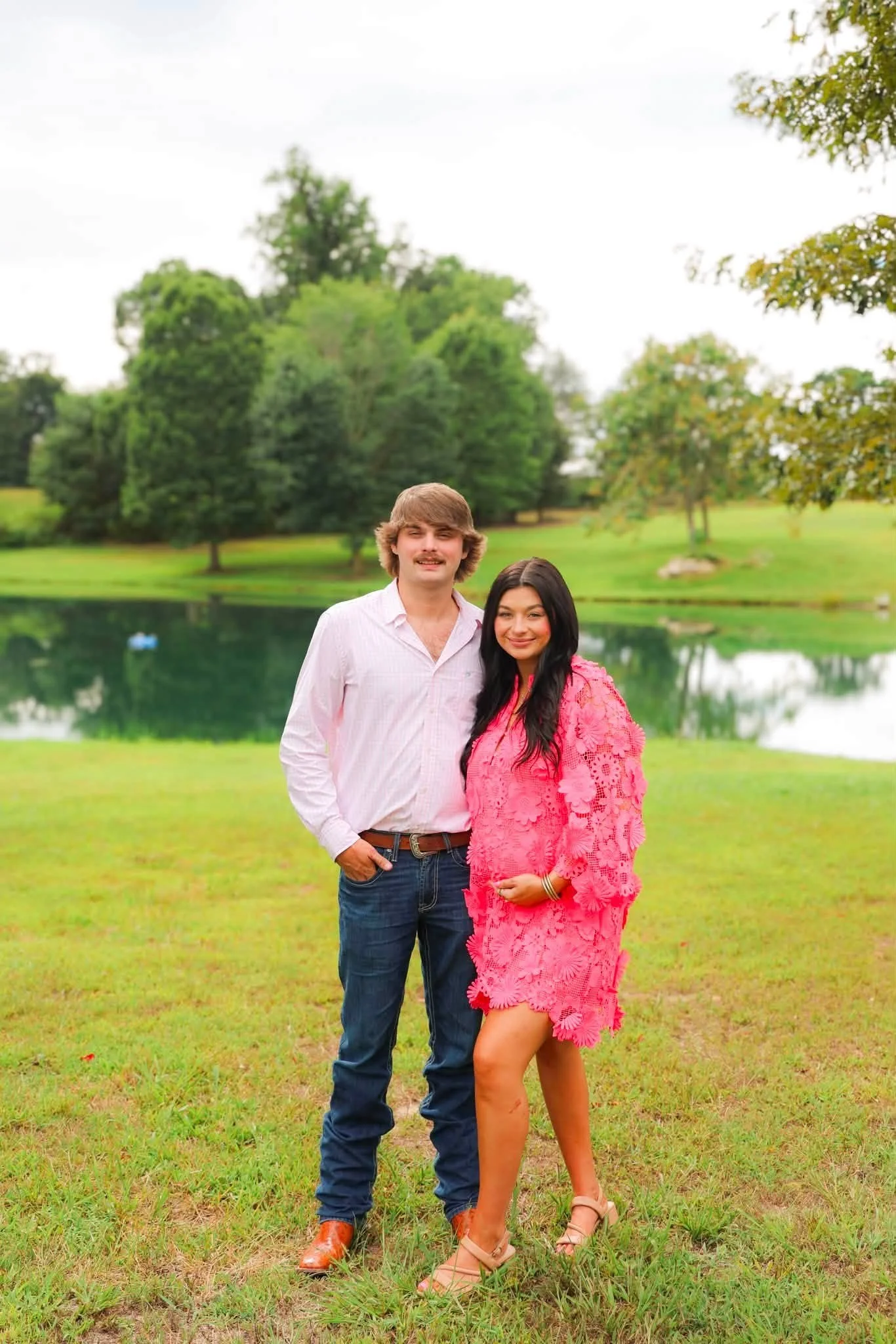 A man and woman standing together outdoors near a lake with trees in the background. The man is wearing a light pink shirt and jeans, and the woman is wearing a pink dress. They are smiling and posing for the photo.