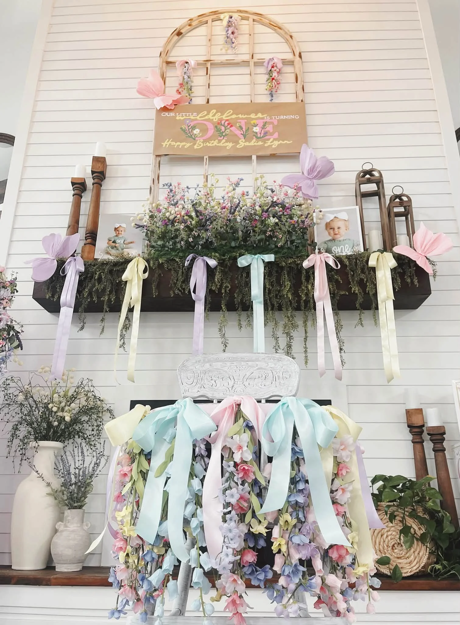 Decorative floral display with pastel ribbons, framed photos of a young girl, and a sign that reads 'Our Little Flower is Turning One, Happy Birthday Sadie Lynn' against a white-paneled wall.