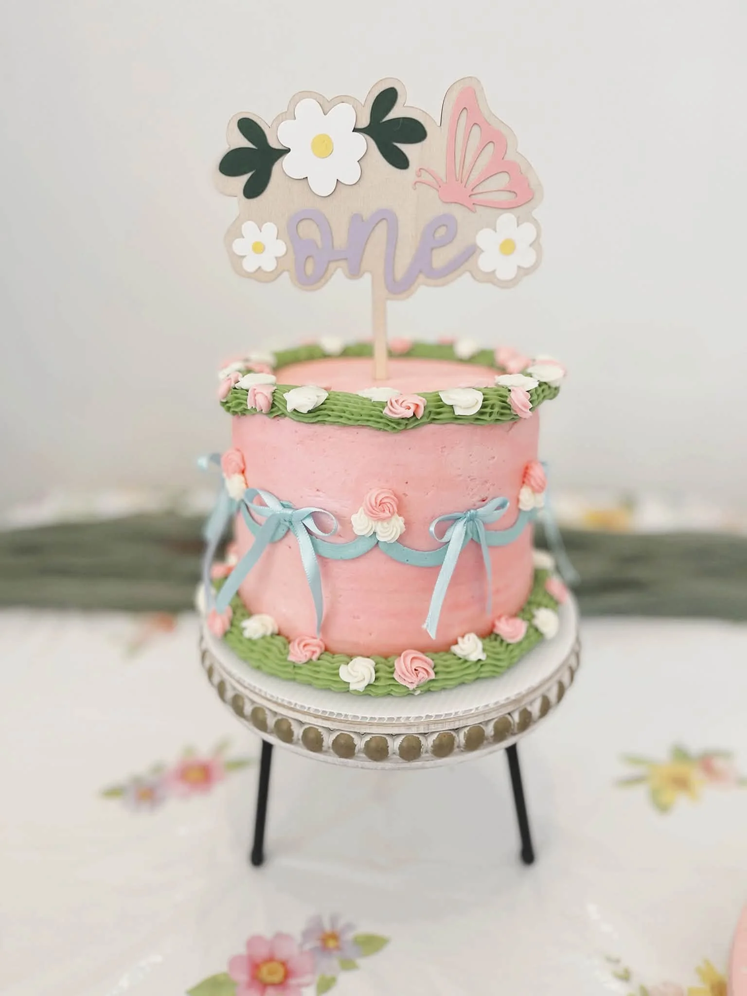 A two-tier pink cake decorated with blue ribbons, white and pink roses, sits on a white cake stand. The top of the cake features a pink butterfly and a sign with the word "LOVE" surrounded by white flowers with yellow centers, and green leaves.