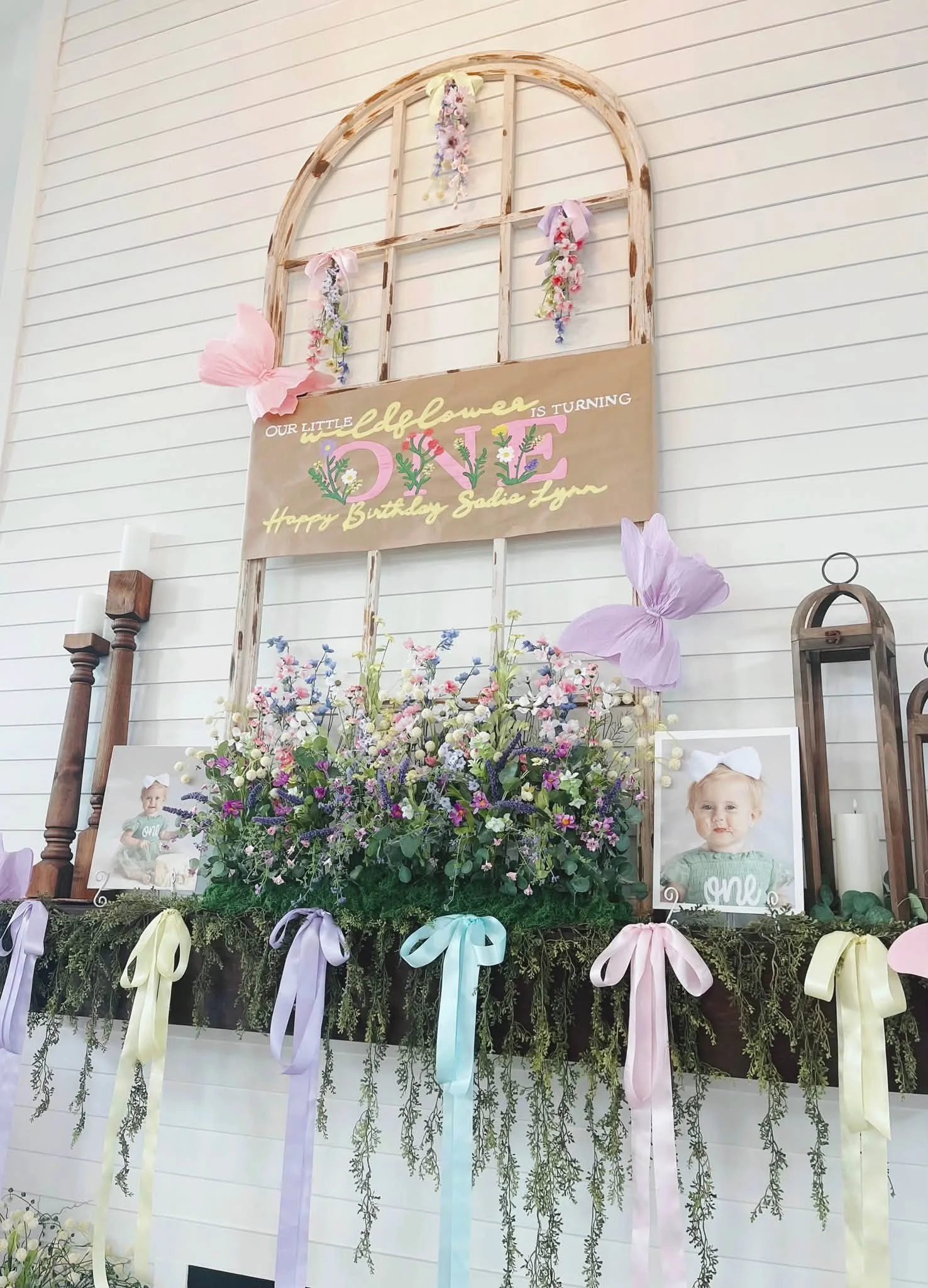 Decorative floral arrangement with framed photos of a young girl, a large floral alphabet spelling 'ONE', and a sign reading 'Our little butterfly is turning 1. Happy Birthday Sadie Faye', with pastel ribbons and greenery.