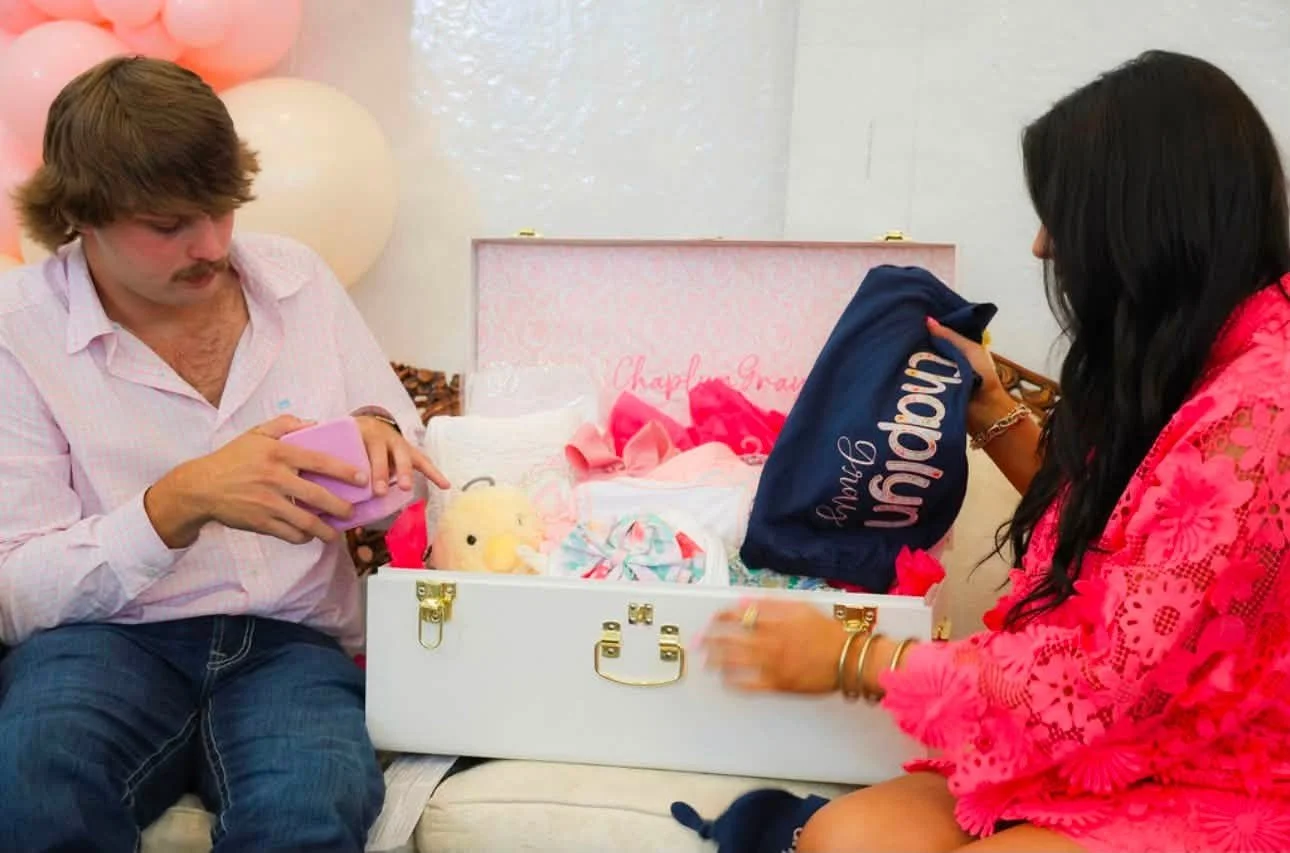 A man and woman opening a baby gift box containing a stuffed toy, baby clothes, and other items, with pink and white balloons in the background.