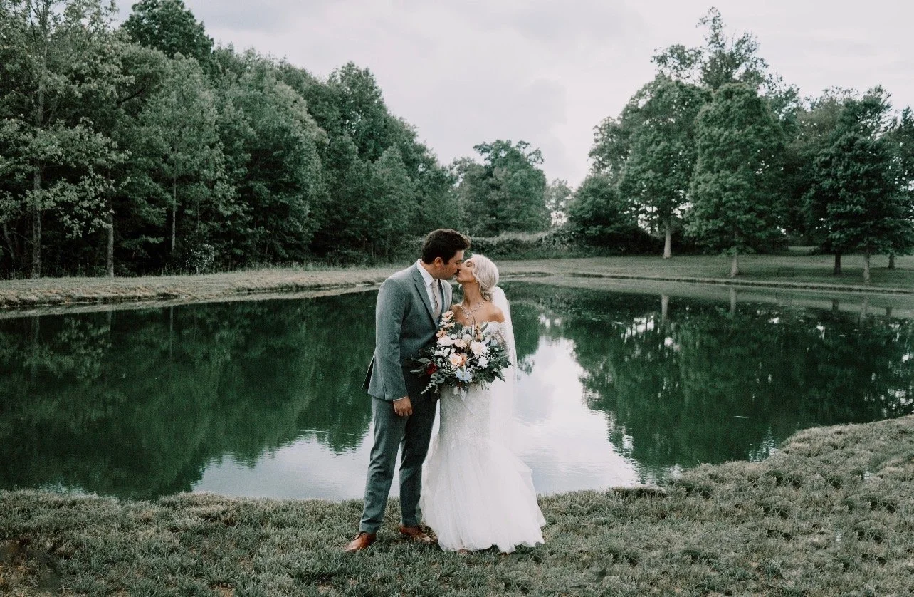 A bride and groom sharing a kiss by a pond with trees in the background.