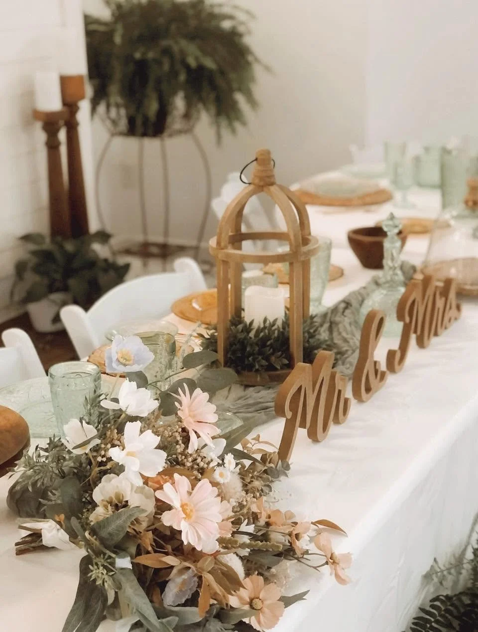 Decorative table centerpiece with a large candle inside a lantern, surrounded by greenery and flowers, with a wooden 'Mr & Mrs' sign, set on a white tablecloth for a wedding or special occasion.