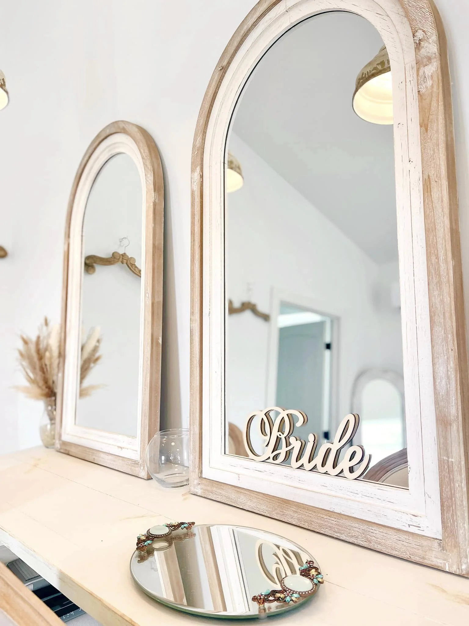 Decorative mirrors with white wooden frames and the word "Bride" inside one mirror, a small glass bowl, and jewelry on a light-colored surface in a room with arrows and hooks on the wall.