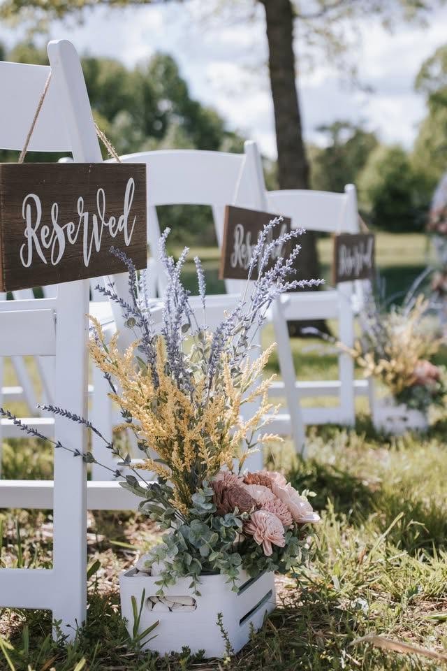 Outdoor wedding seating area with white chairs, reserved signs, and floral arrangements in a white crate.