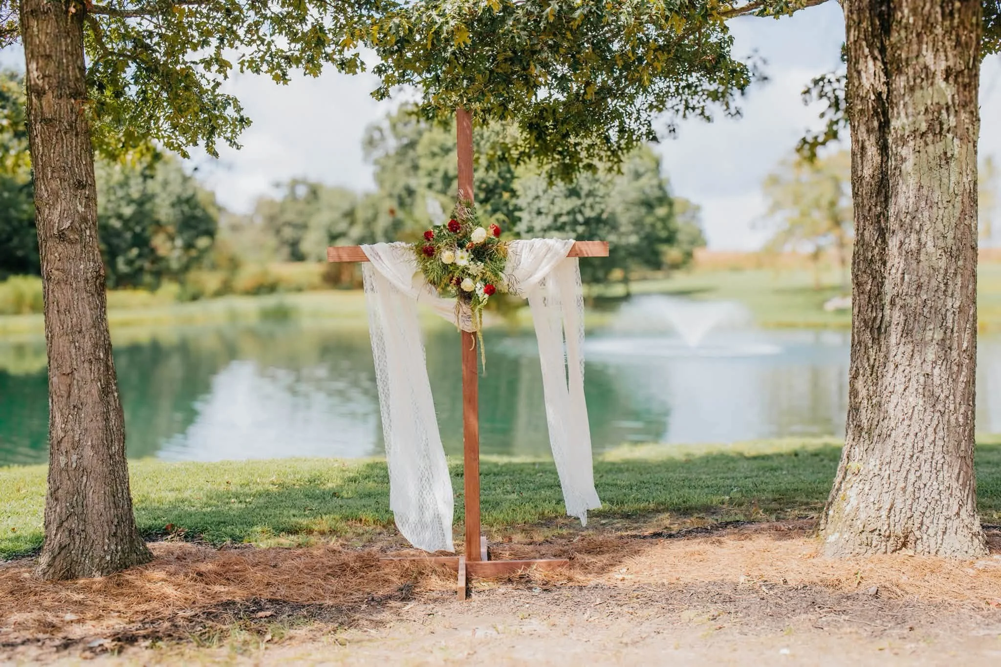 Outdoor wedding altar decorated with a floral arrangement of red and white flowers, with white fabric hangs on a wooden cross in front of two trees by a lake.