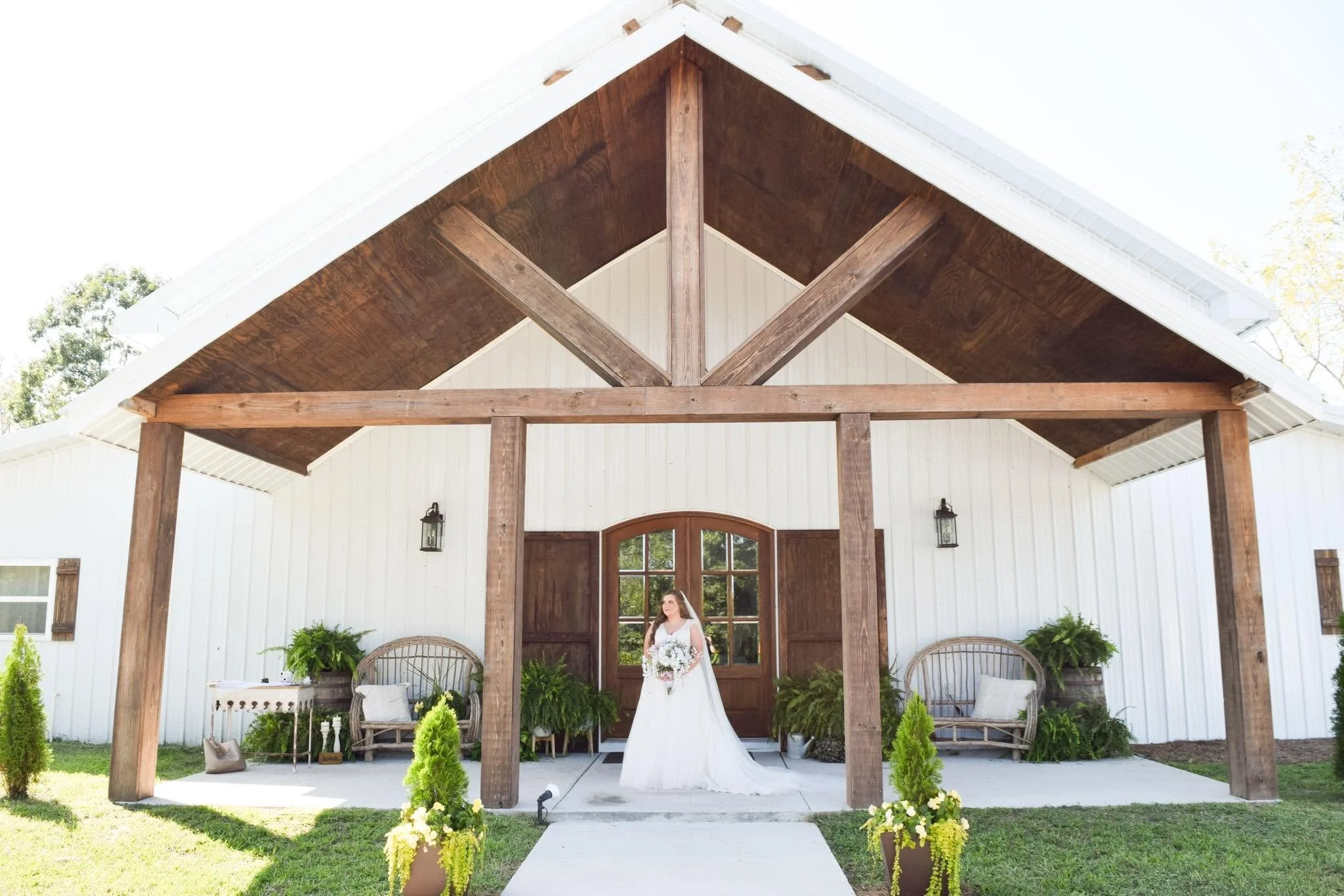 A woman in a wedding dress holding a bouquet standing outside a rustic barn with white walls and wooden beams under the overexposed sky.