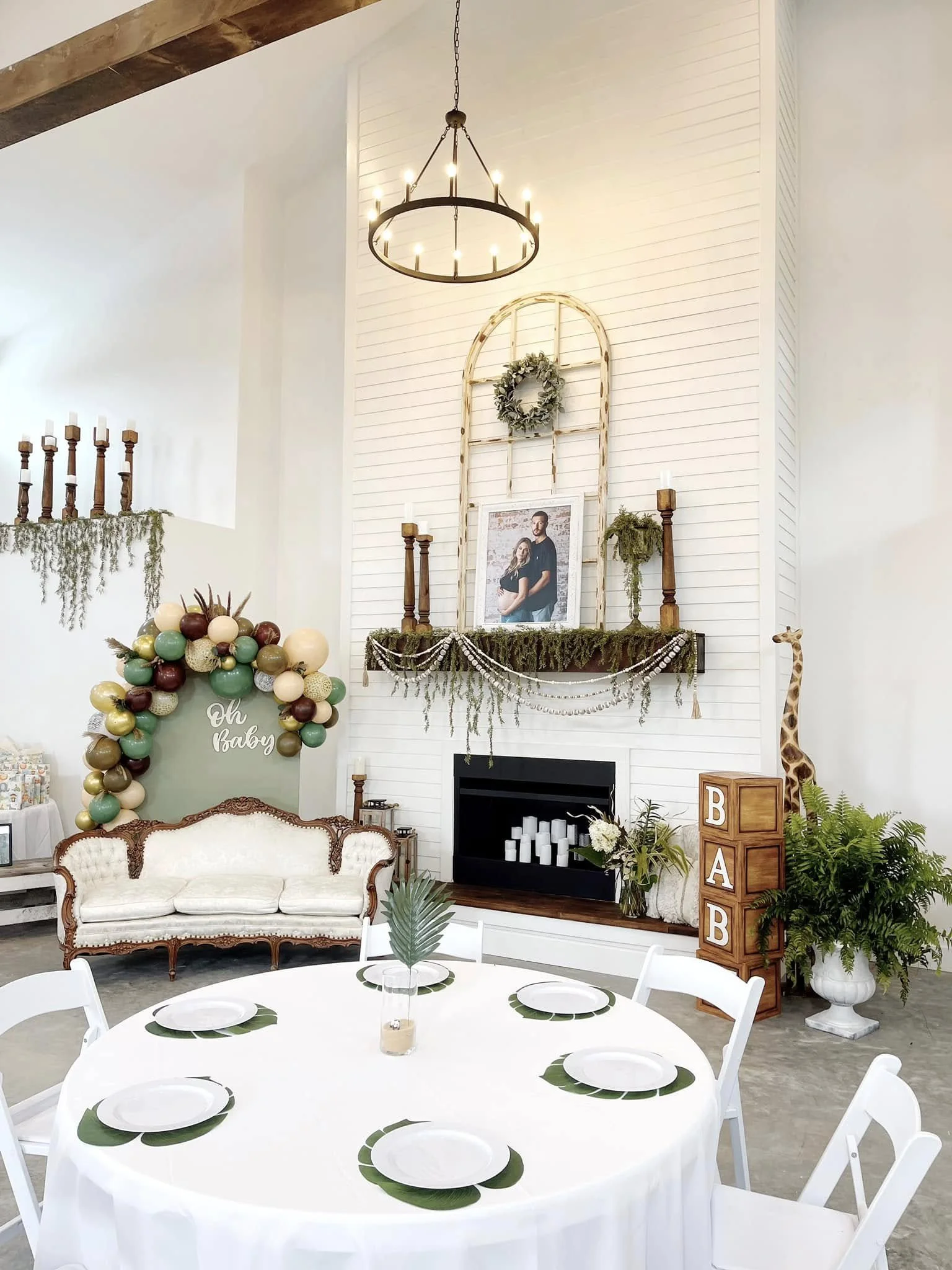 Decorative indoor space for a celebration, featuring a white ornate sofa, a round table with white tablecloth and green leaf-themed plates, a balloon arrangement with shades of green, gold, and burgundy, a white shiplap wall decorated with a frame and greenery, candles, a framed couple's photo, a shelf with candles and plants, a wooden pillar with blocks spelling 'BABY', a mural of a giraffe, a large wreath, and a wooden chandelier with candles.
