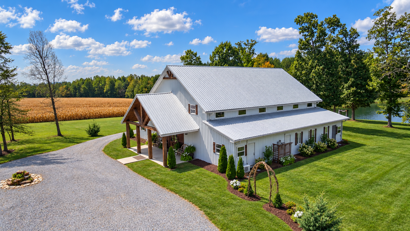 A white farmhouse with a metal roof, surrounded by a well-maintained lawn, trees, and a gravel driveway, with a body of water and a field of crops in the background under a partly cloudy sky.