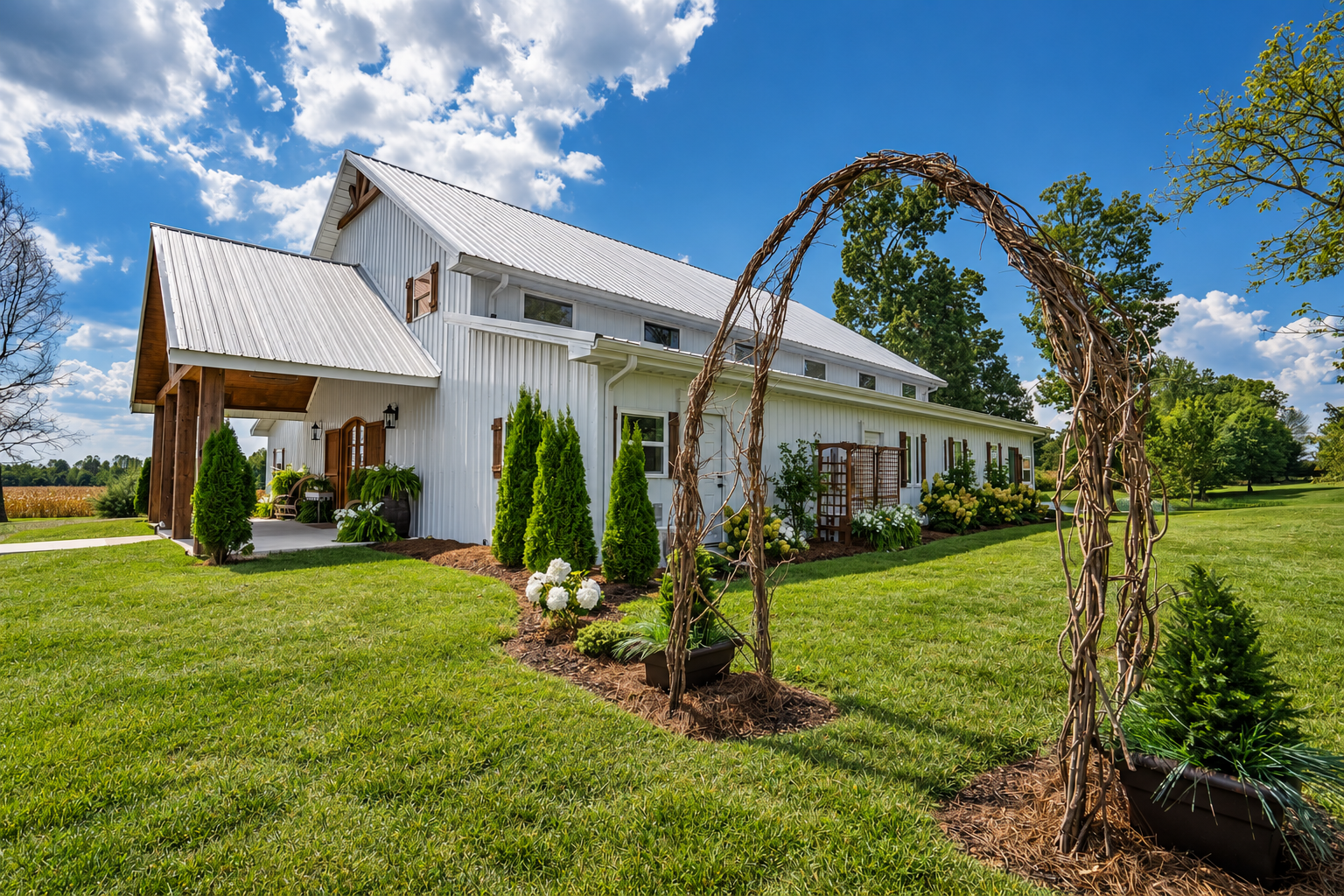 A white barn-style house with a metal roof surrounded by a green lawn, plants, and flowers, under a blue sky with clouds, with a twisted vine arch in the foreground.