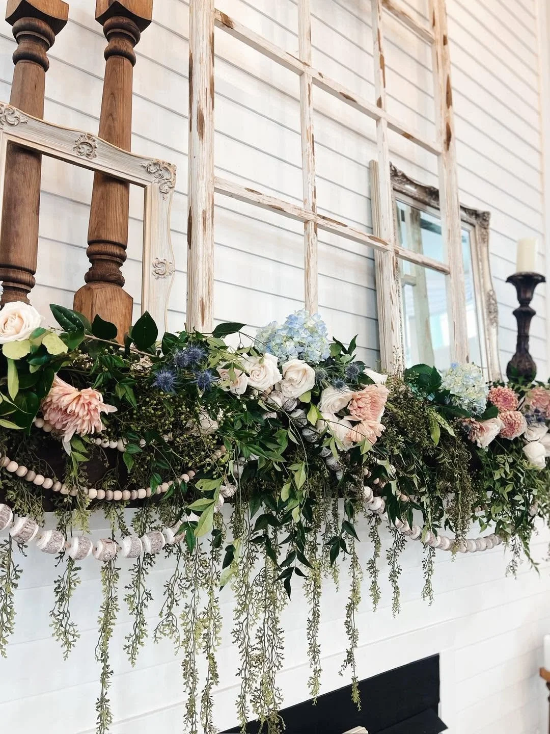 White wooden wall decorated with floral arrangement including white roses, pink flowers, blue hydrangeas, and greenery, with empty vintage frames and a candlestick on either side.