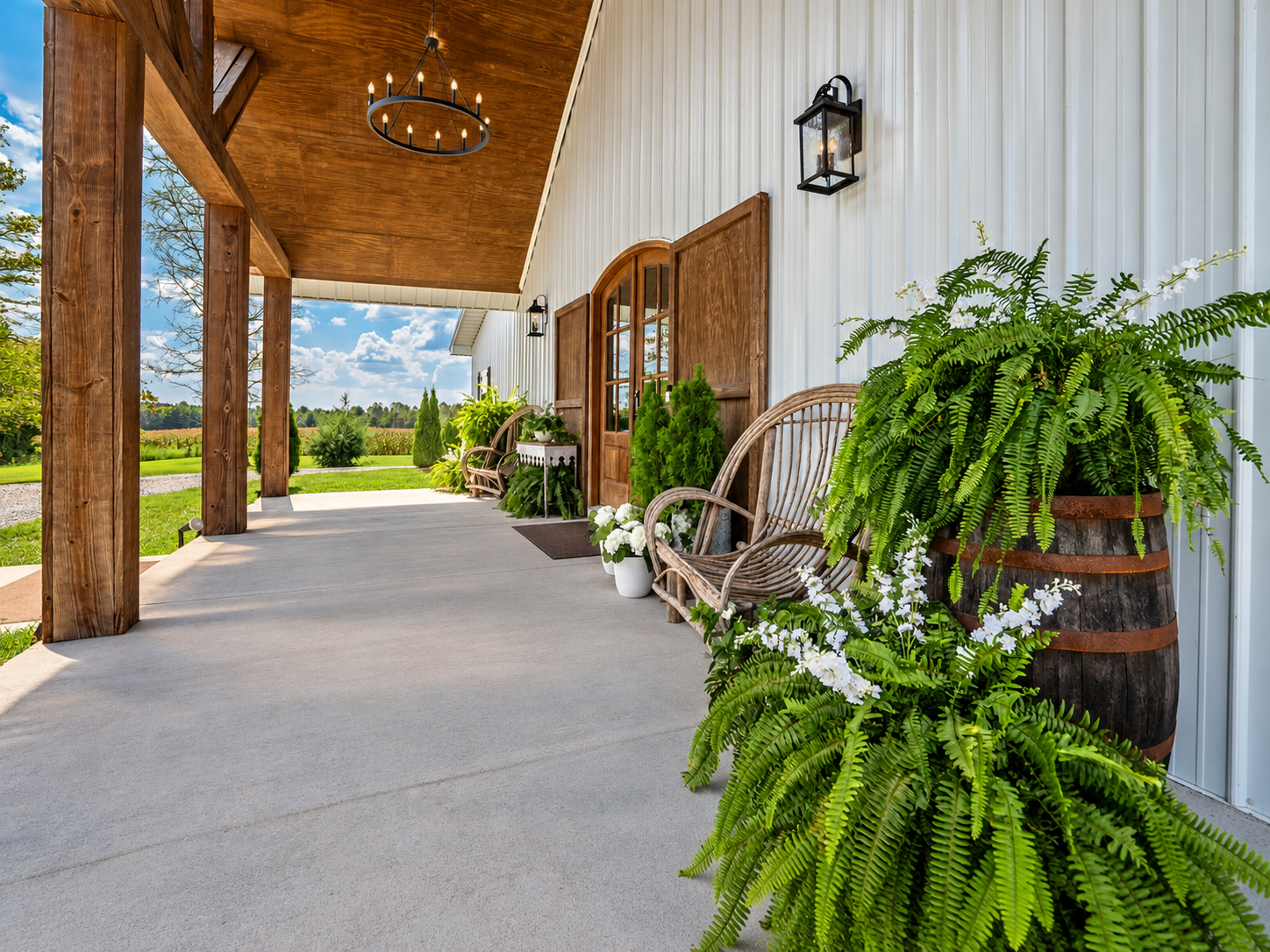 Front porch of a white farmhouse with wooden accents, potted plants, outdoor wicker chairs, and a scenic rural landscape in the background.