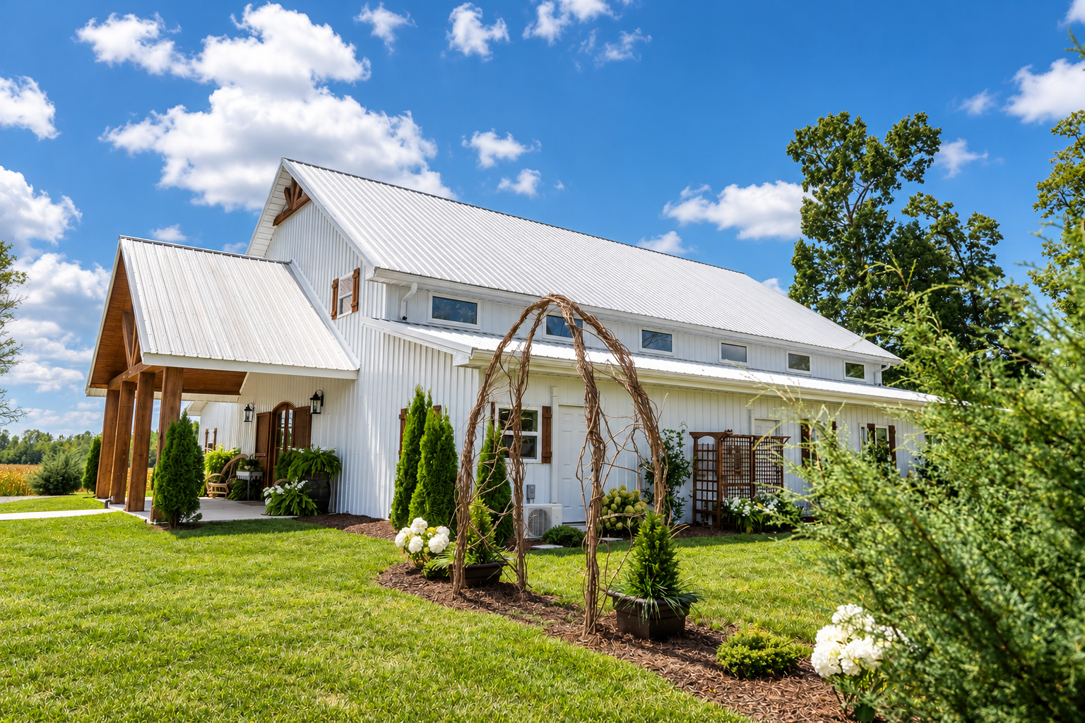 White farmhouse-style house with a metal roof, surrounded by a lush green lawn, trees, and plants, under a partly cloudy sky.