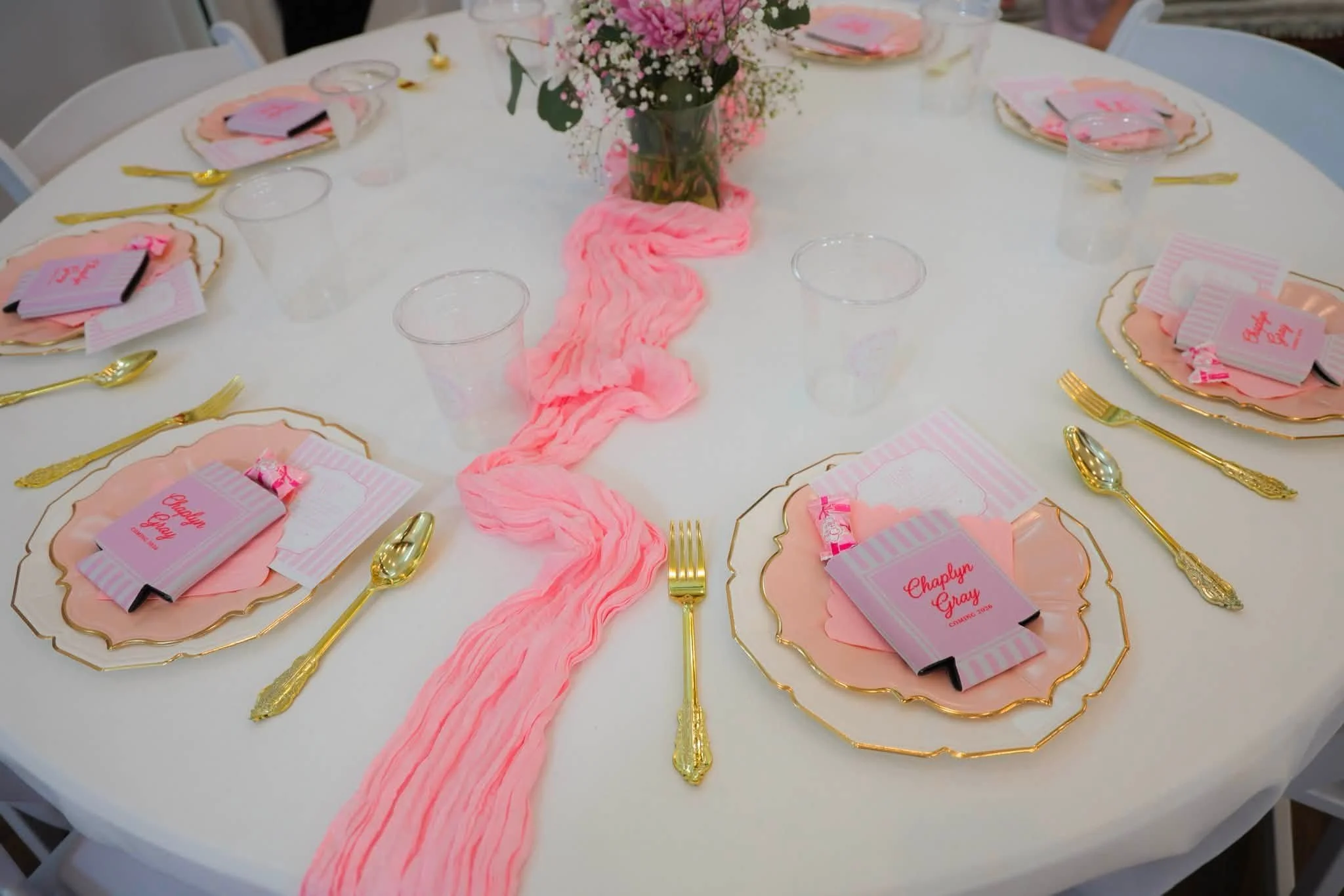 Round table decorated with pink and gold accents, floral centerpiece with pink and white flowers, pink streamer runner, place settings with gold utensils, pink and white placemats, and small pink and white favor cards.