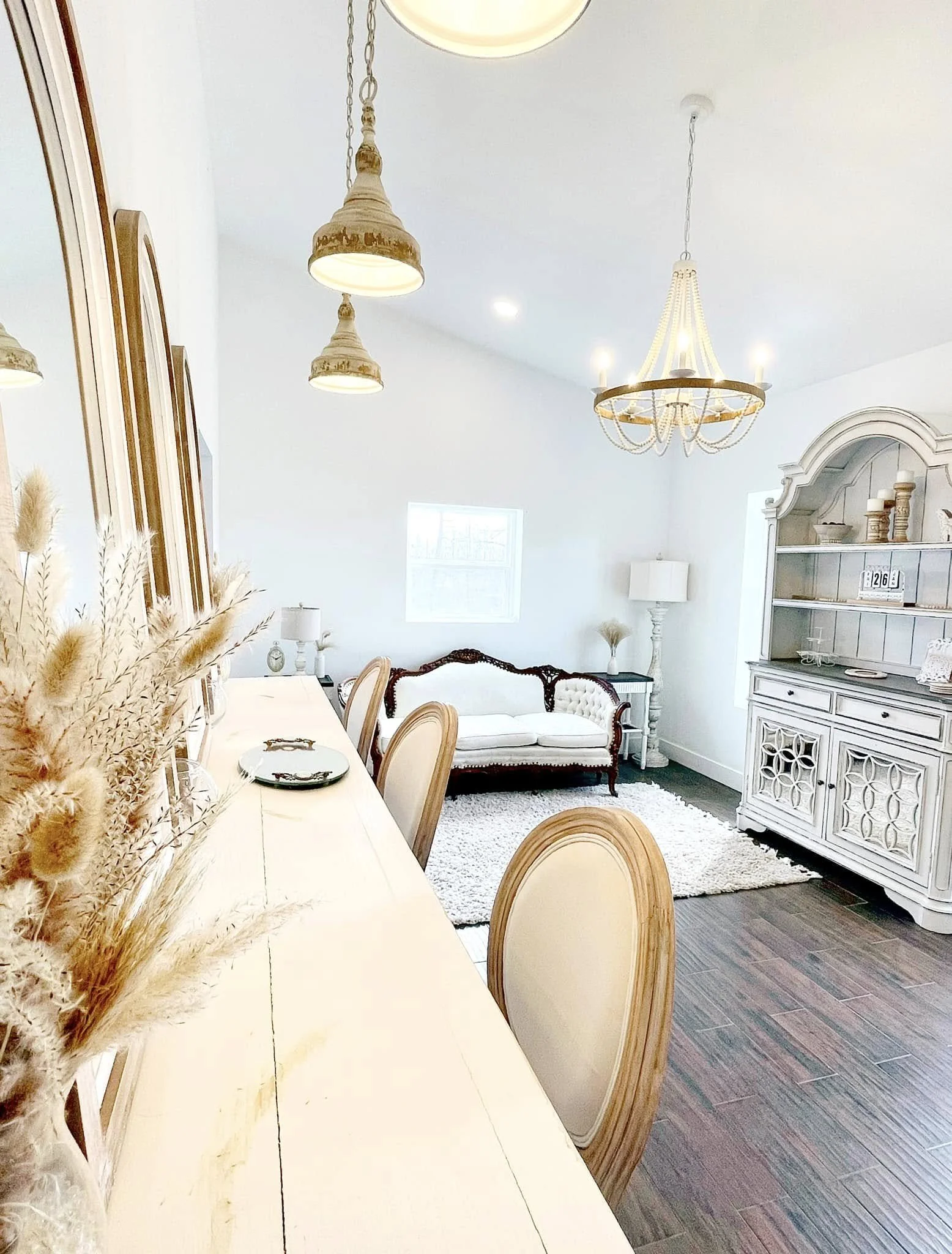 The image depicts a bright, elegantly decorated living room with a white color scheme. There is a long dining table with wooden chairs, a white ornate sofa, and a white cabinet with decorative items. The ceiling features a modern chandelier and penda