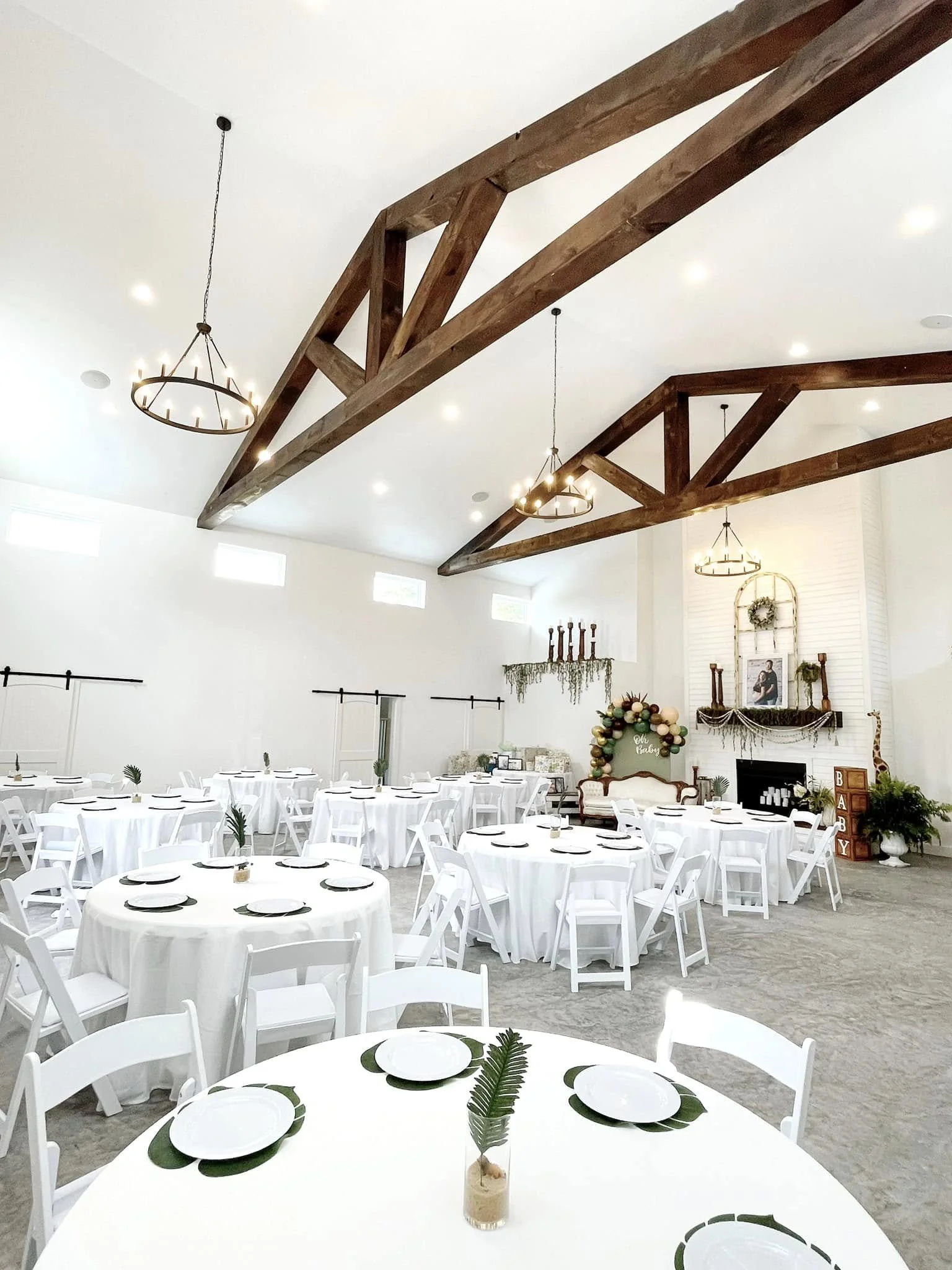 Indoor event space decorated for a celebration with white tablecloths, white chairs, and simple centerpieces featuring a leaf in a small glass jar. The room has high ceilings with wooden beams and chandelier lighting, white walls, and a decorated fire mantel with a wreath and framed photograph.