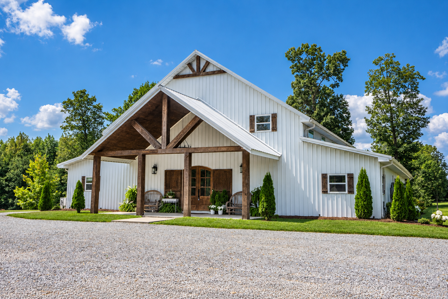 A modern farmhouse style home with white siding and a wooden front porch, surrounded by lush green trees and a gravel driveway under a blue sky with scattered clouds.