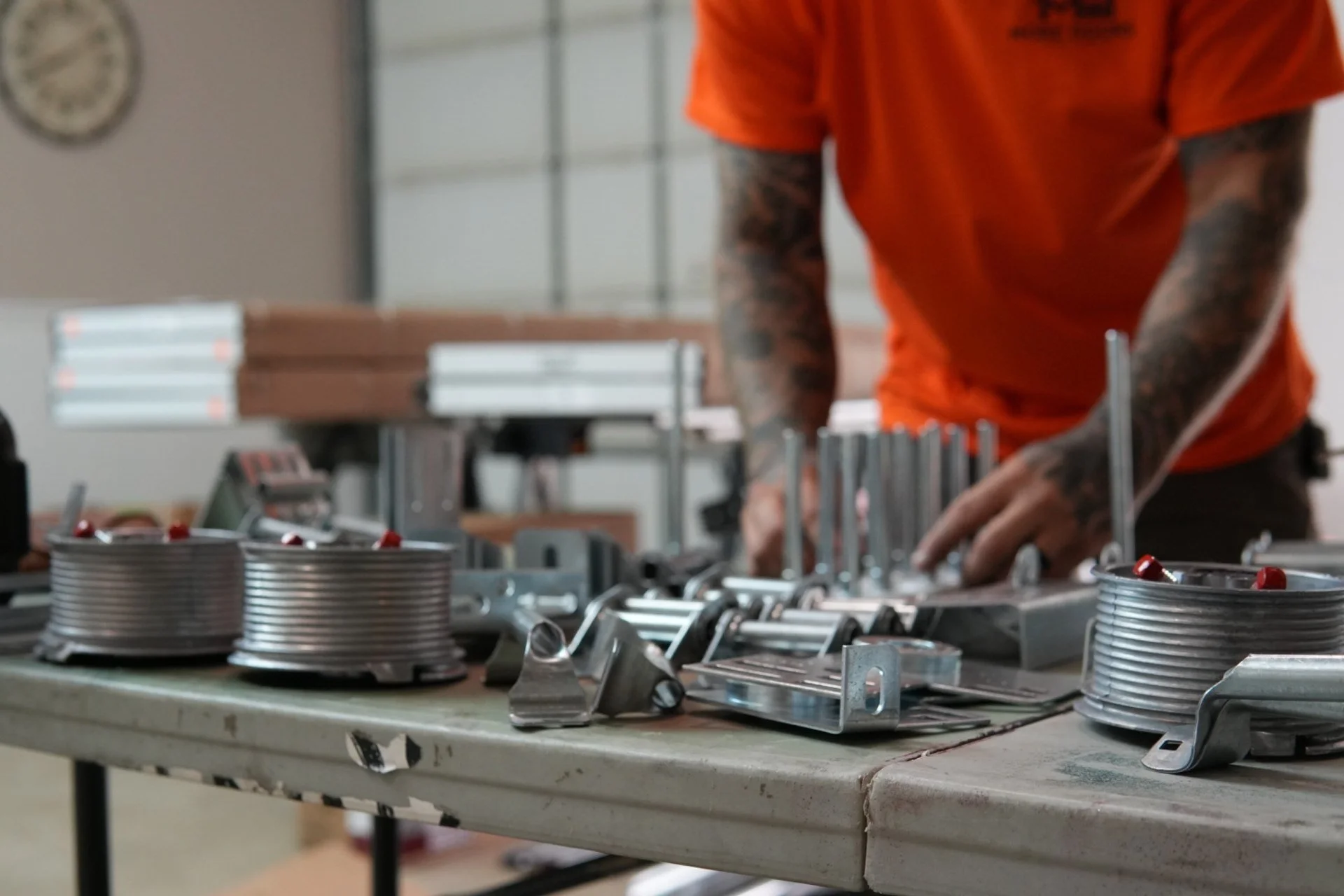 A worker in an orange uniform working on a mechanical assembly line with metal components, with cans and tools on a workbench in the foreground.