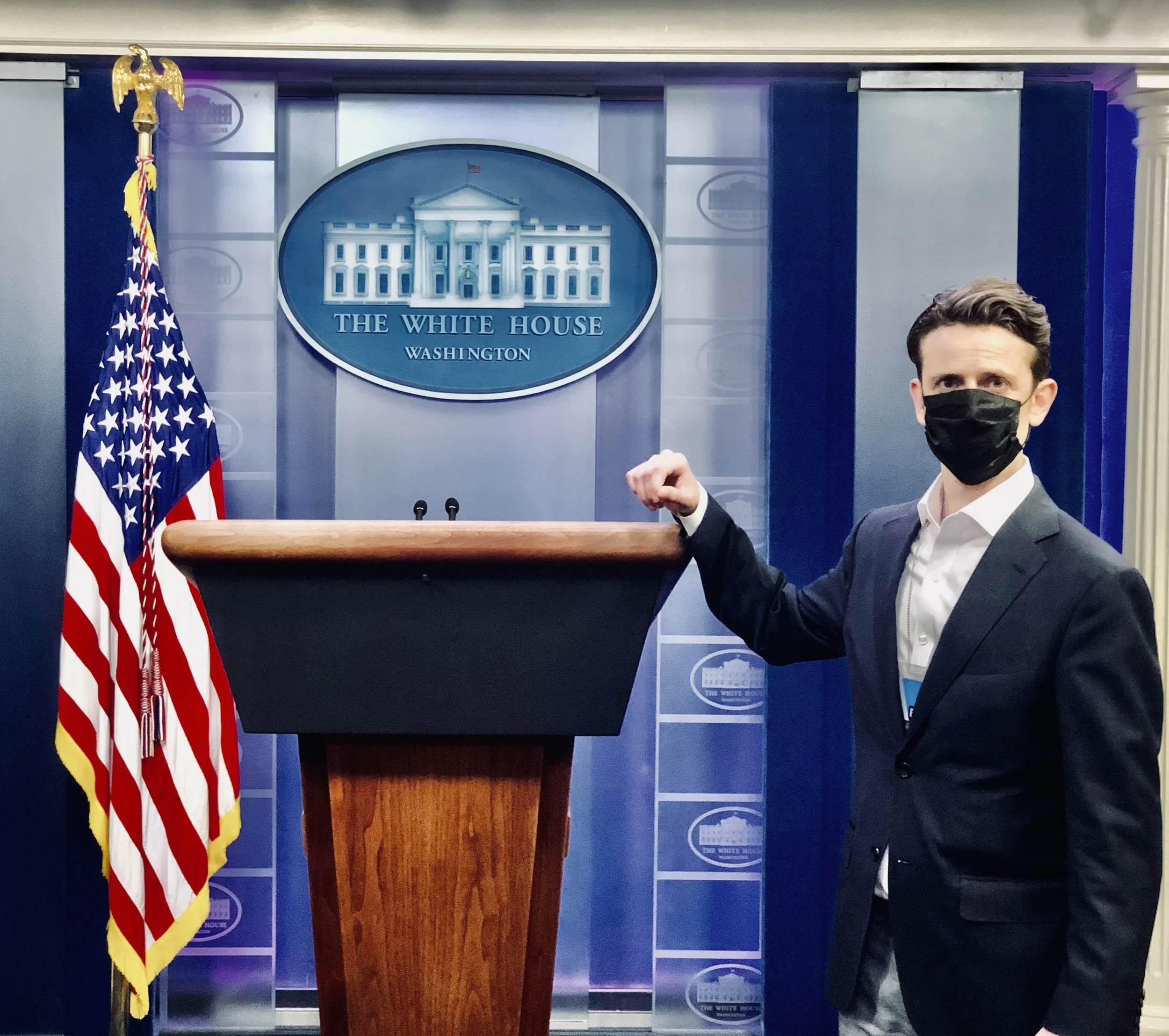 Press briefing room at the White House with a podium, American flag, and the White House logo on the backdrop. A person wearing a black mask and suit stands near the podium, raising their right fist.