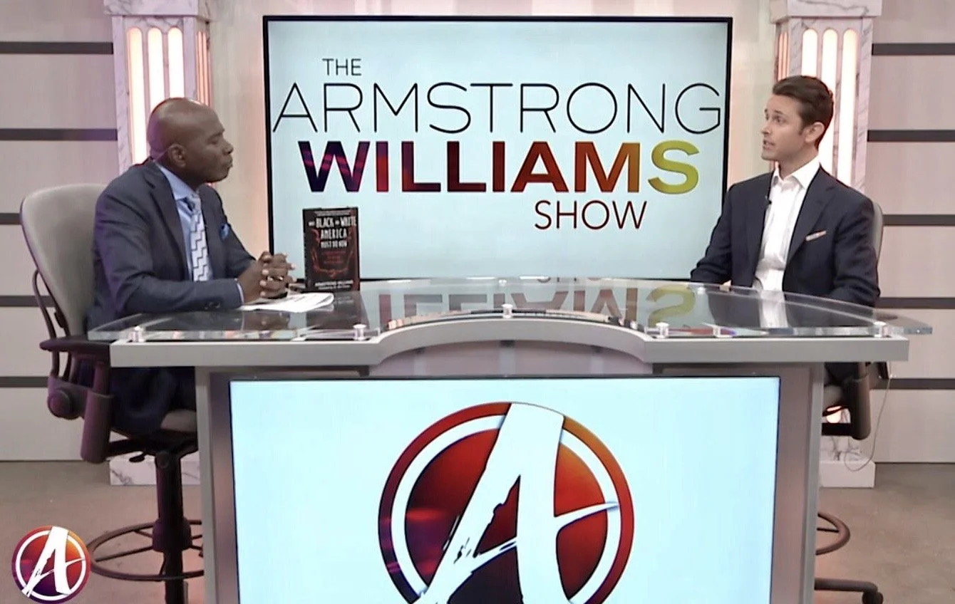 Two men are seated at a news desk in a television studio. Behind them is a large screen with the text "The Armstrong Williams Show" and a book displayed on the desk. The desk has a large logo with a stylized letter "A" on the front.