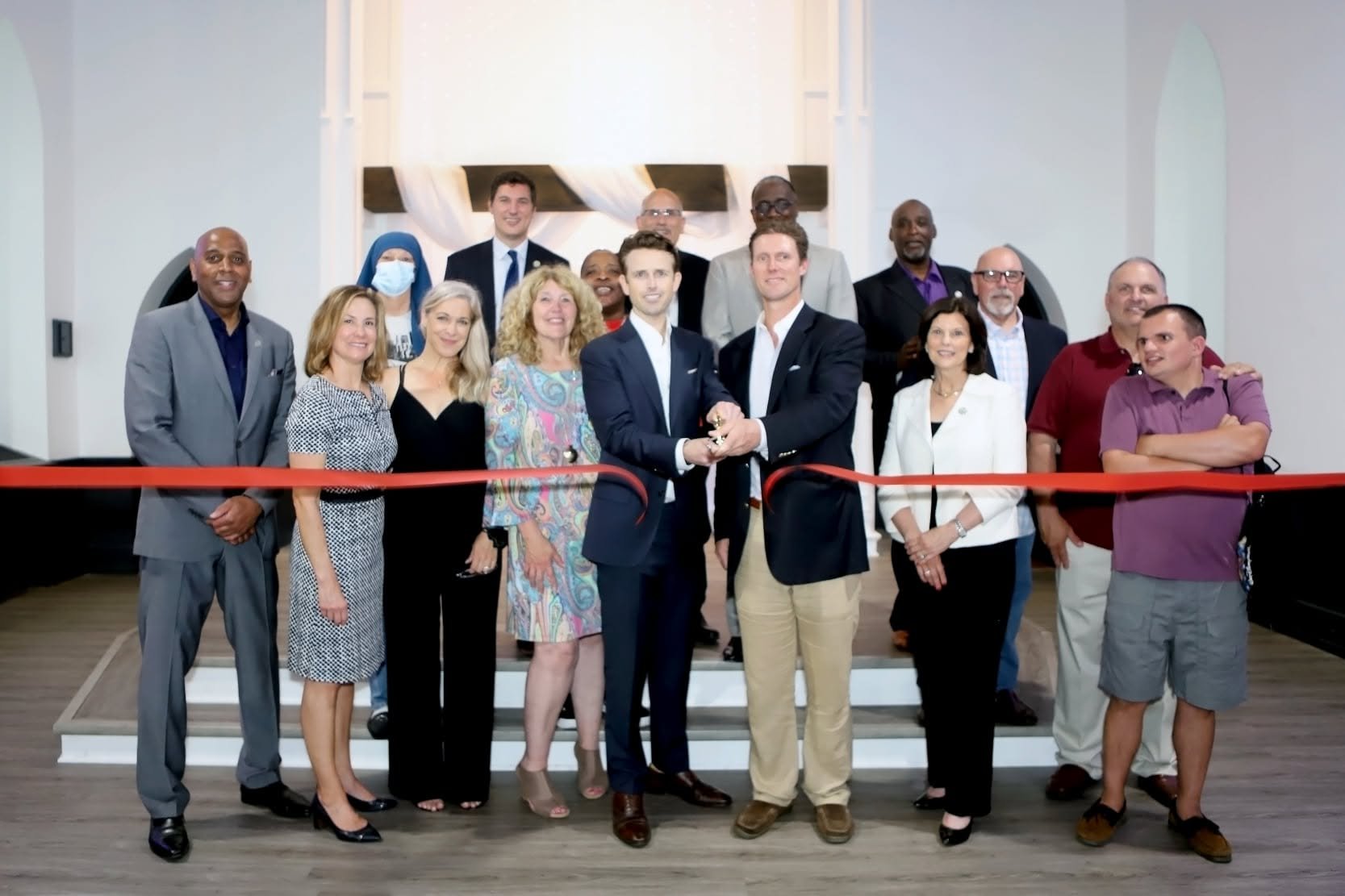 Group of diverse adults at a ribbon-cutting ceremony in an indoor setting, standing on stairs with a red ribbon in front of them.