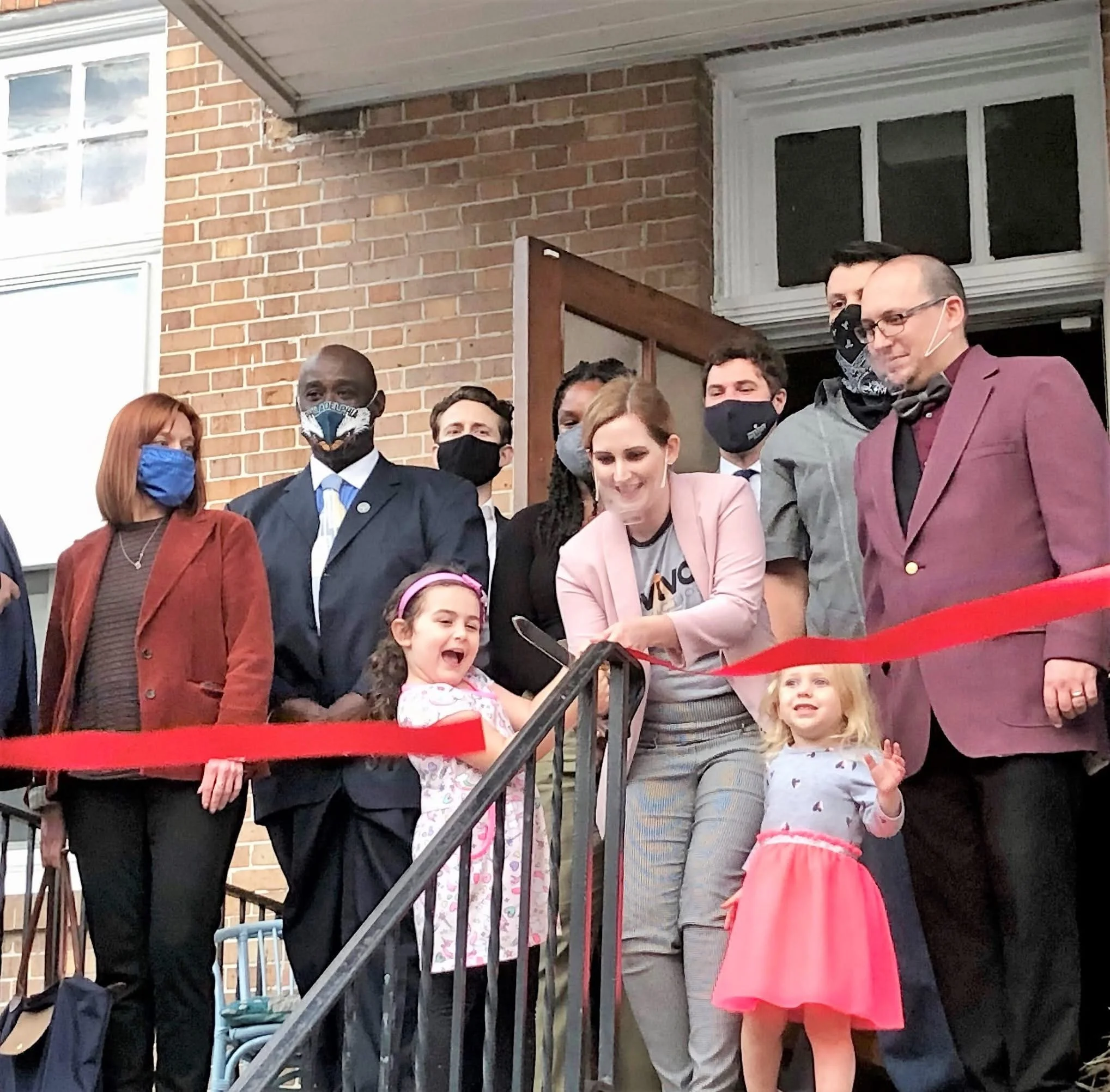 Group of people gathered outside near a brick building. Some are wearing face masks. A young girl in a pink dress and white jacket is in the front, smiling and holding a book.