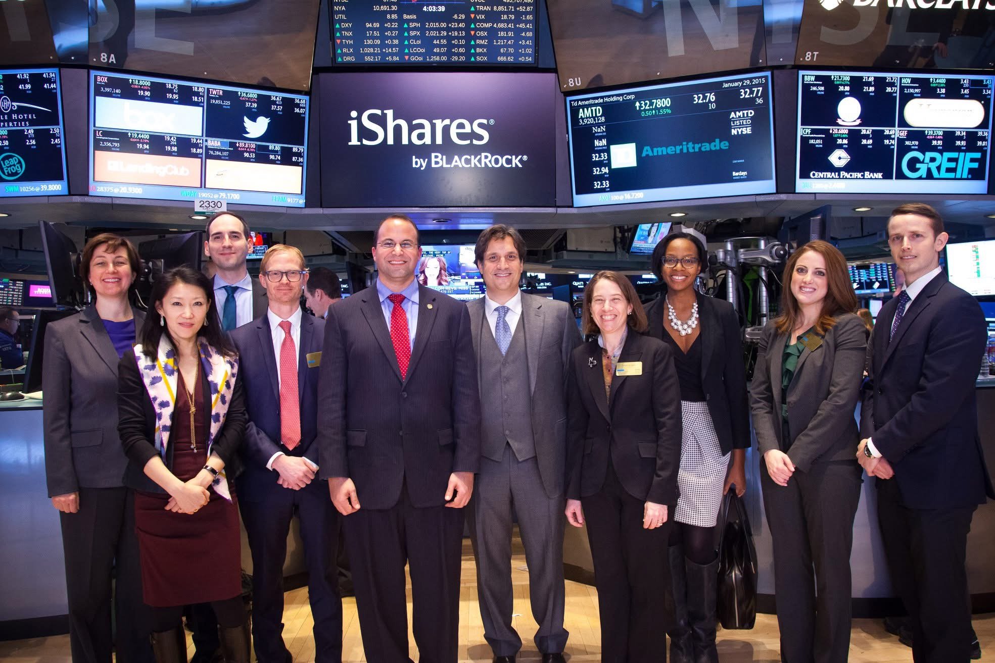 Group of nine diverse business professionals, four women and five men, standing together on a trading floor with multiple screens displaying stock market data in the background.
