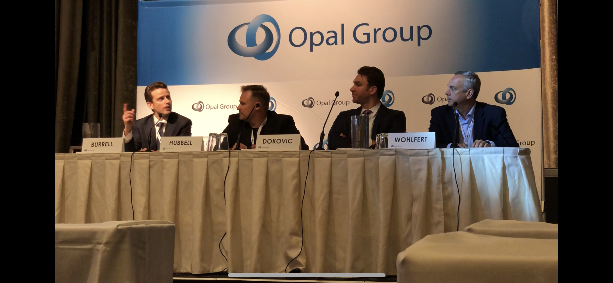 A panel of four men sitting at a long table during a conference, with microphones and nameplates that read Burrell, Hubbell, Djokovic, and Wohlert. Behind them is a large backdrop with the Opal Group logo and name.