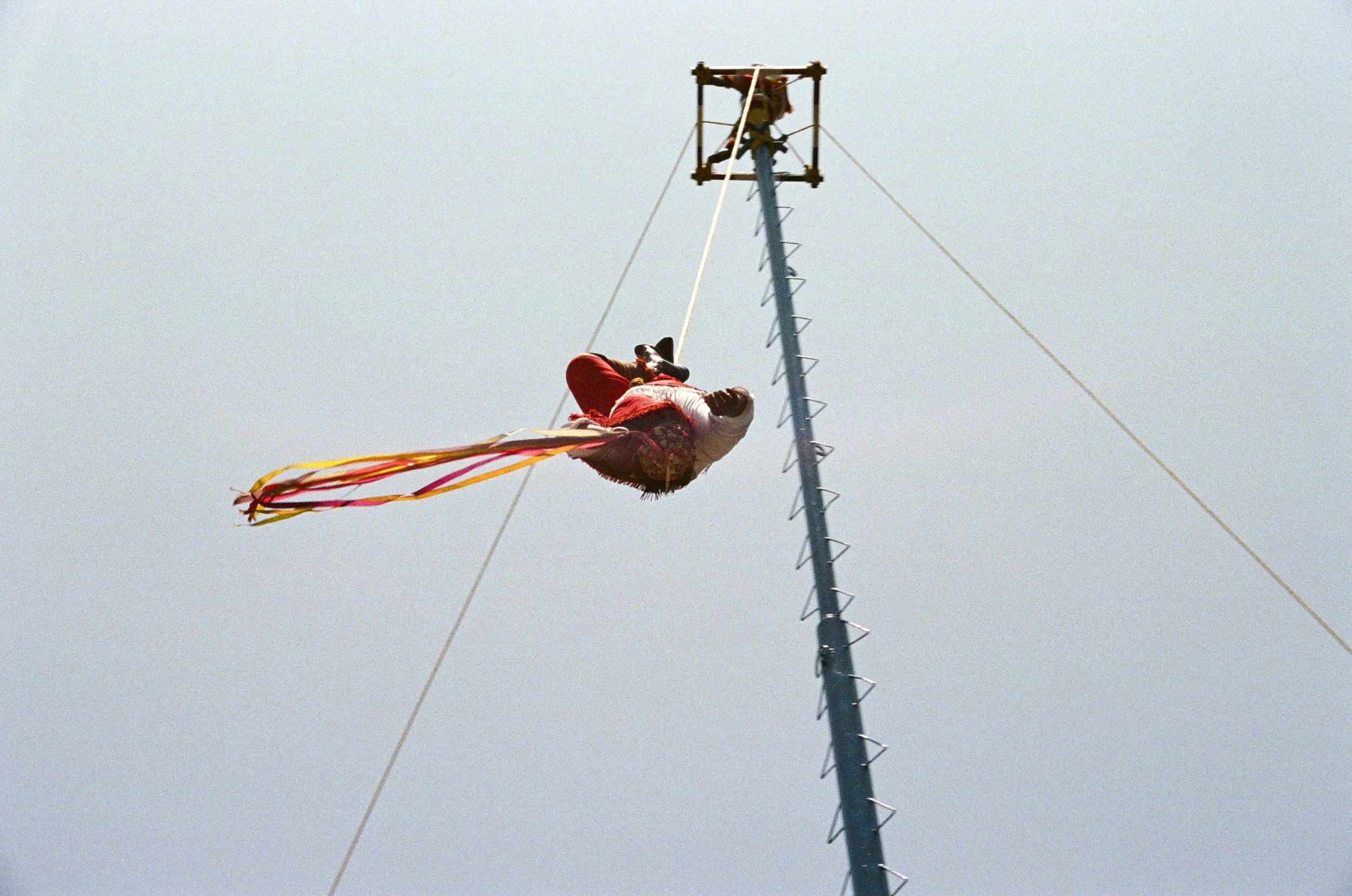 A person hanging upside down from a tall pole with colorful ribbons attached to their waist, with a blue sky in the background.