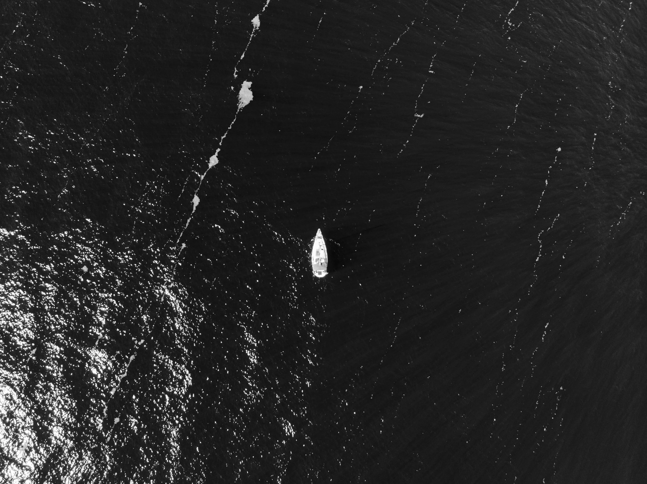 Aerial view of a boat sailing in open water, creating white wake streaks behind it. The image is in black and white, with sunlight reflecting off the water's surface.