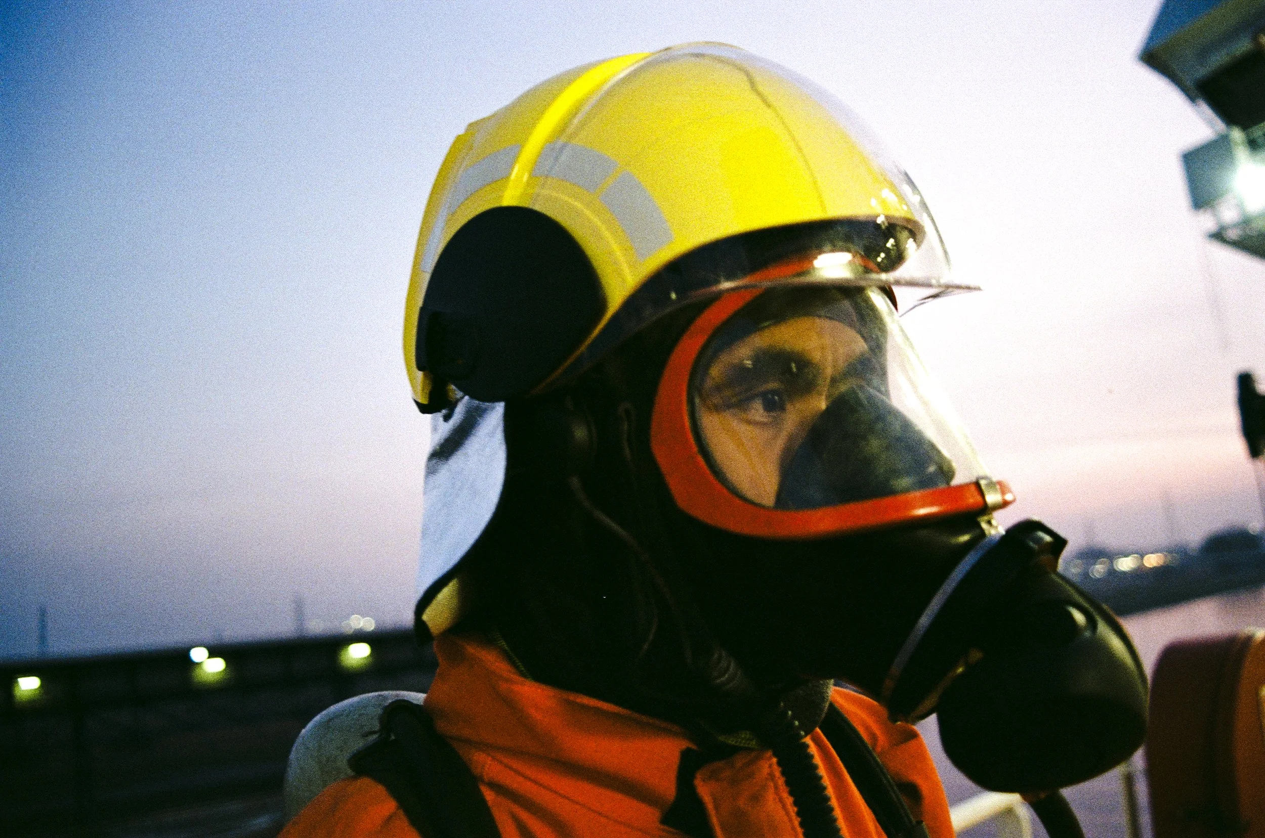 A person wearing a yellow safety helmet, protective goggles, and an orange jacket, with a dark face mask, is outdoors at dusk or dawn, possibly riding a motorcycle or scooter.