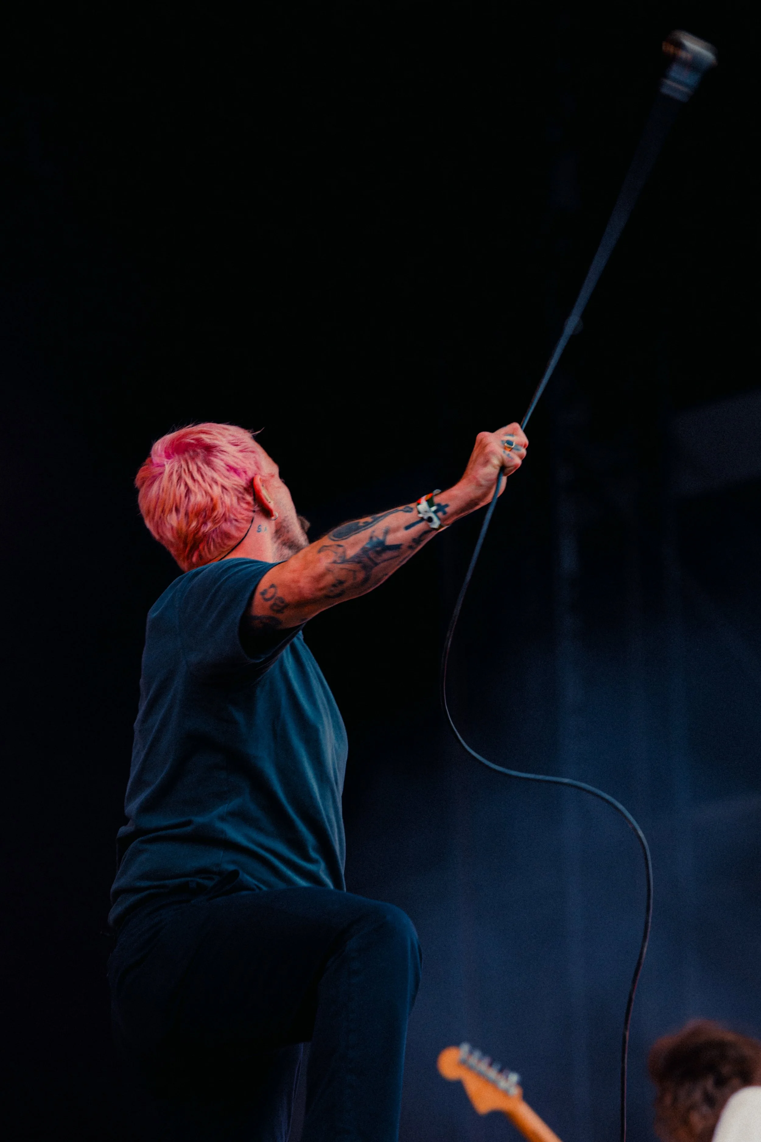 A performer with pink hair on stage holding a microphone stand in their right hand, wearing a dark t-shirt and tattoos on their right arm, with a guitar player in the background.