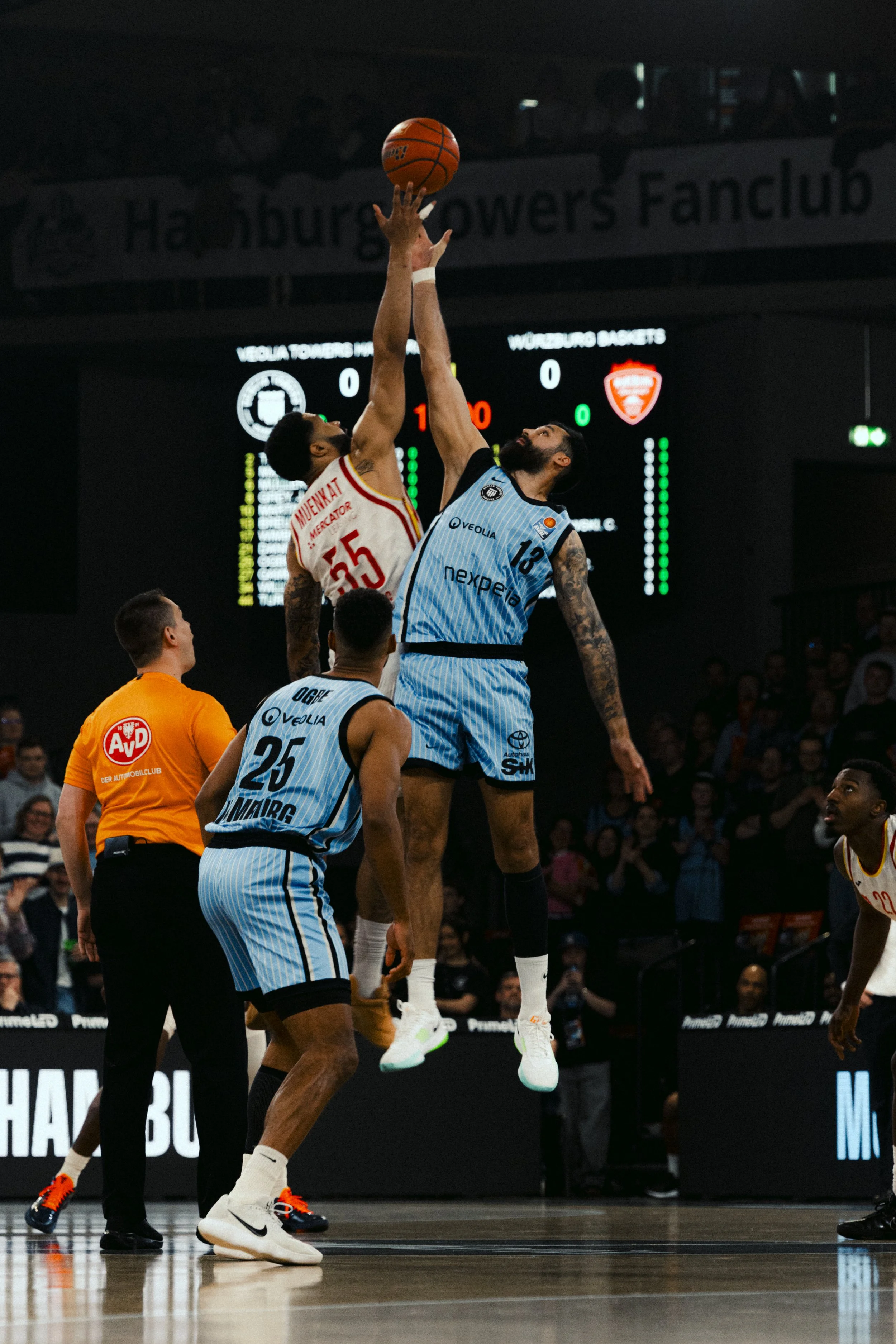 Two basketball players are jumping in the air, competing for the ball during a game. One player is wearing a light blue uniform with number 13, and the other is in a white uniform with number 35. An official in an orange shirt and black pants is watc