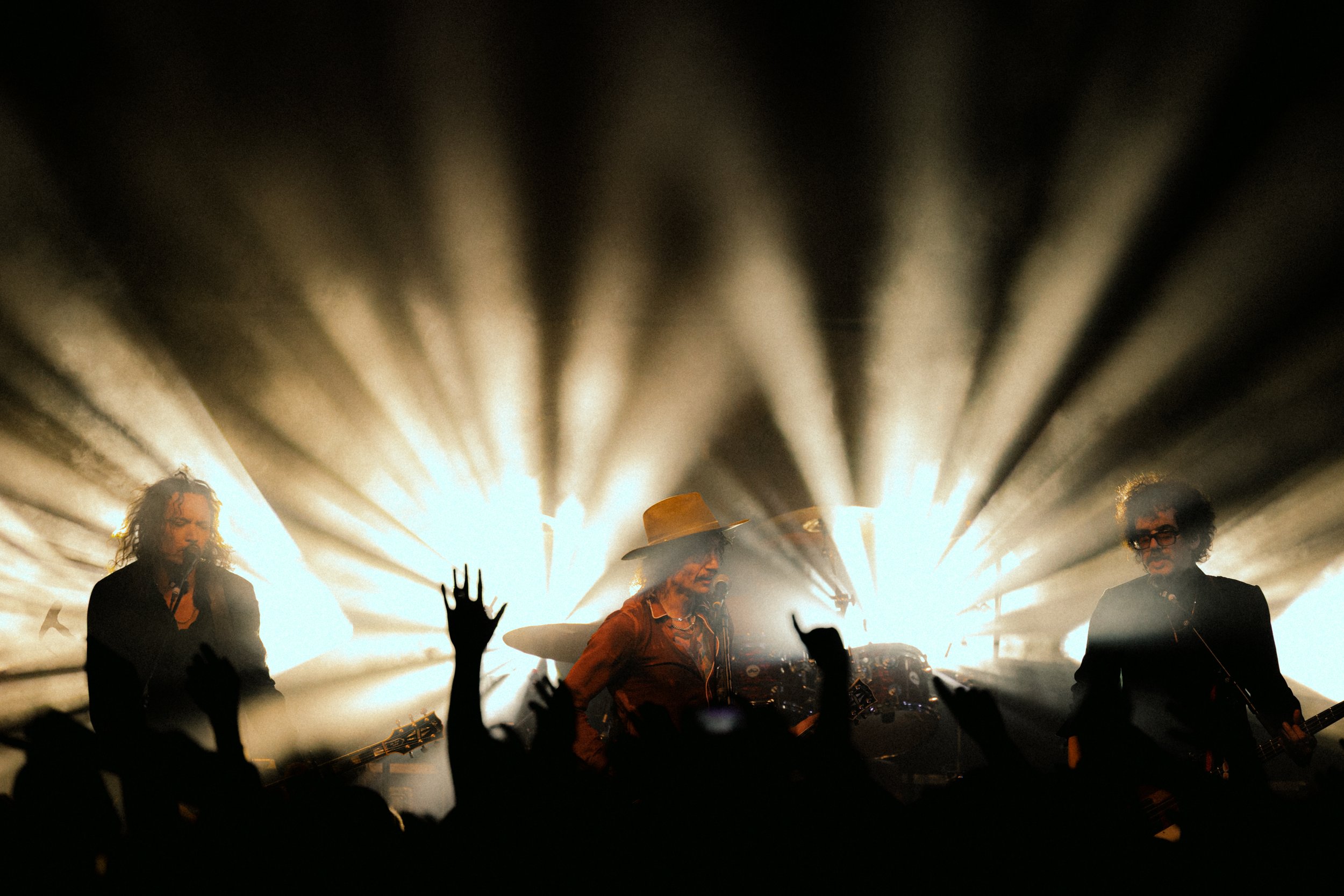 Musicians performing on stage with bright backlighting creating a silhouette effect, and audience members raising their hands.