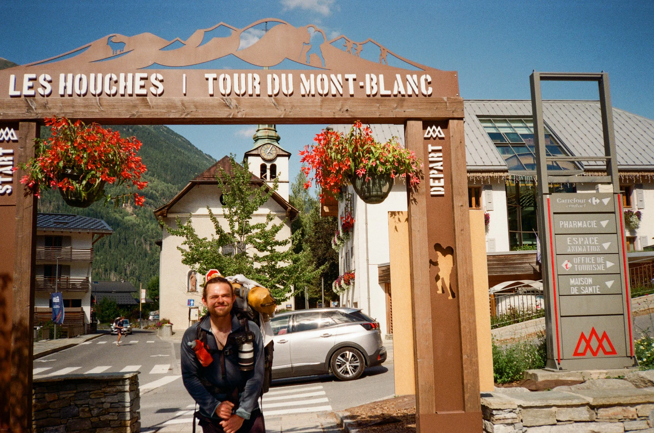A man with a beard, wearing a dark jacket, backpack, and carrying a camera, stands in front of an entrance sign that reads 'Les Houches | Tour du Mont-Blanc.' Behind him, there are hanging flower baskets, a small white church with a clock, residential and commercial buildings, and a mountain landscape with a clear blue sky.