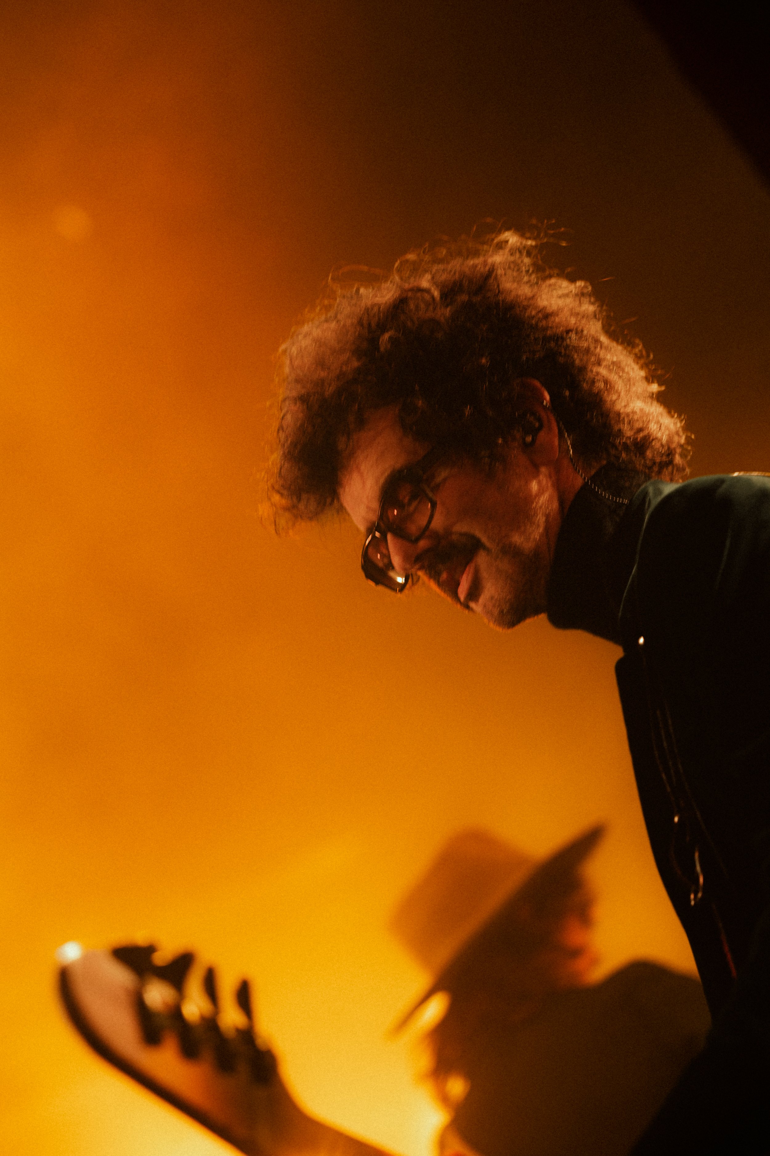 A man with curly hair and glasses, wearing a chain earring, is looking down. The background is yellow and blurred with some dark shapes and a graphic of a dinosaur in the bottom left corner.
