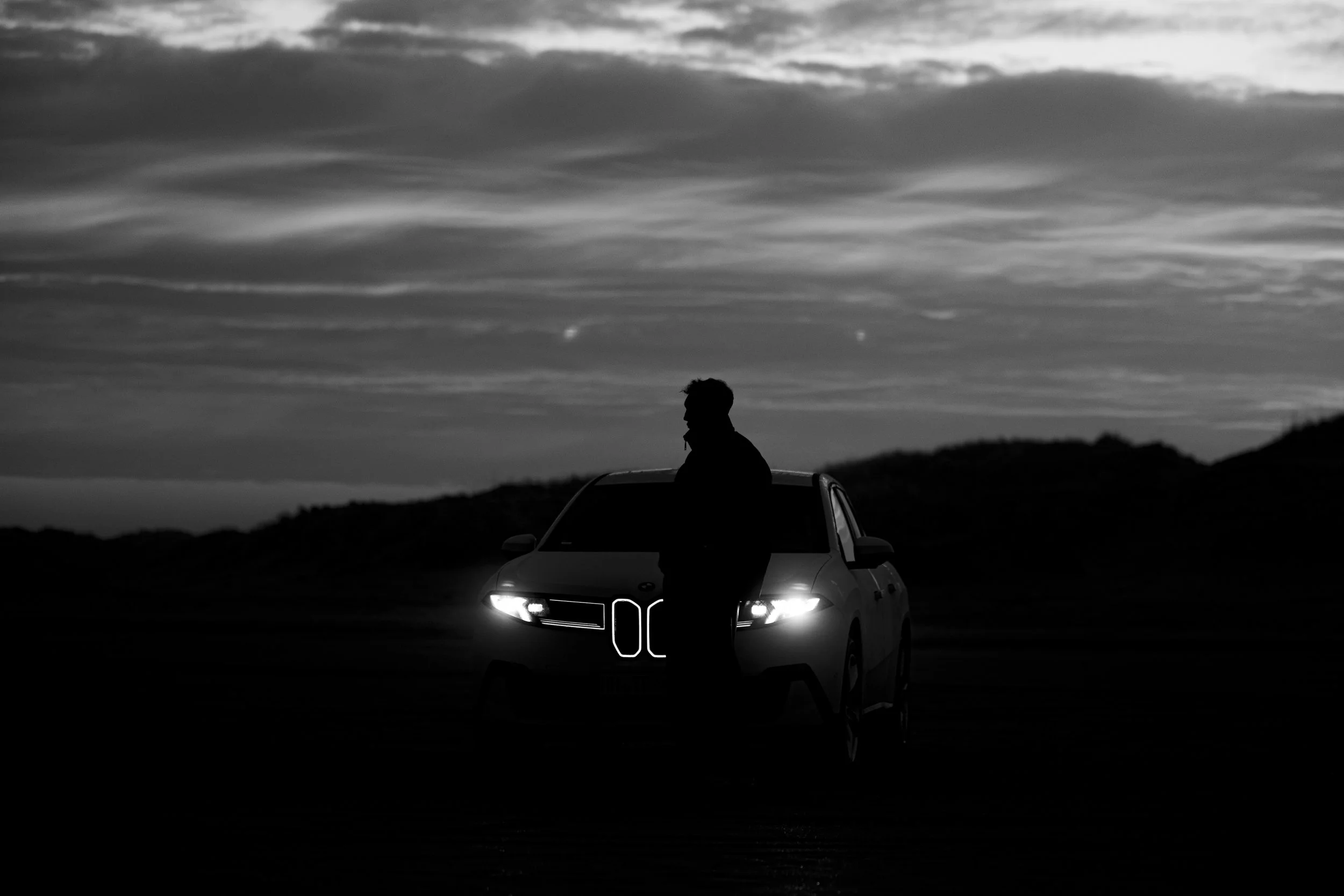 A person standing next to a car with headlights on, against a backdrop of a cloudy sky at dusk or dawn.