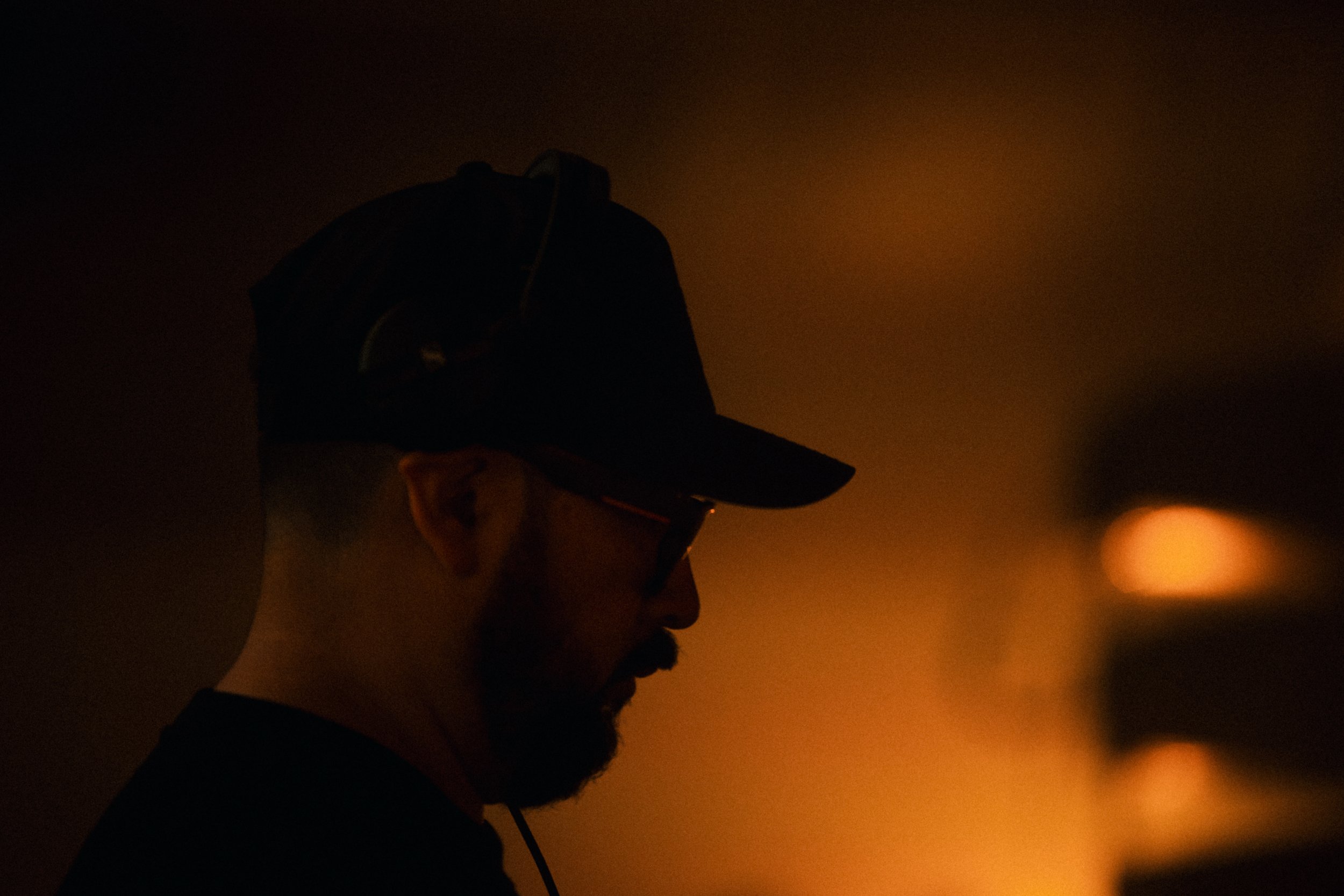 A person wearing glasses and a cap, with a dark background and warm lighting.