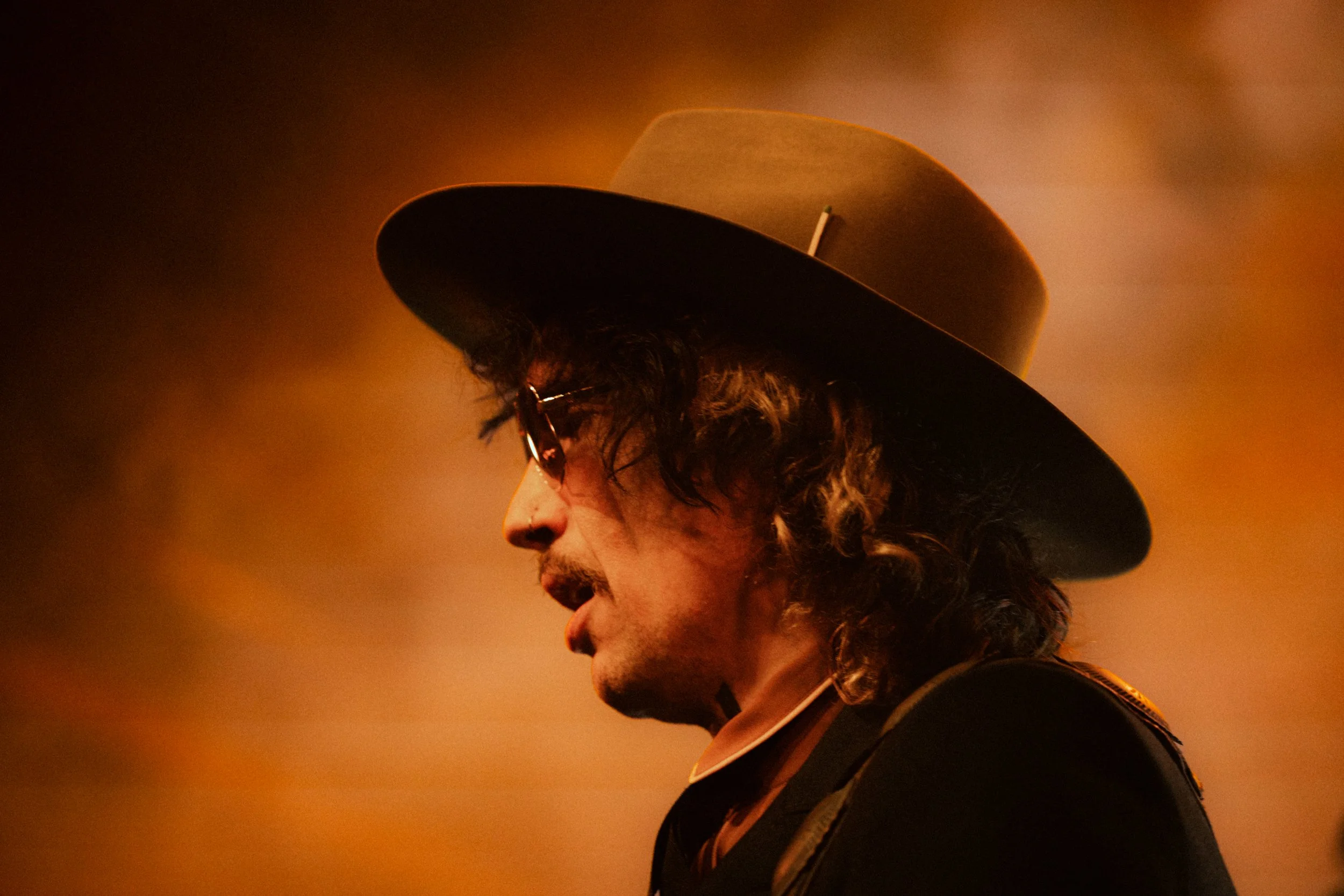 A man with curly hair wearing a fedora and glasses, looking to the side, with warm lighting.