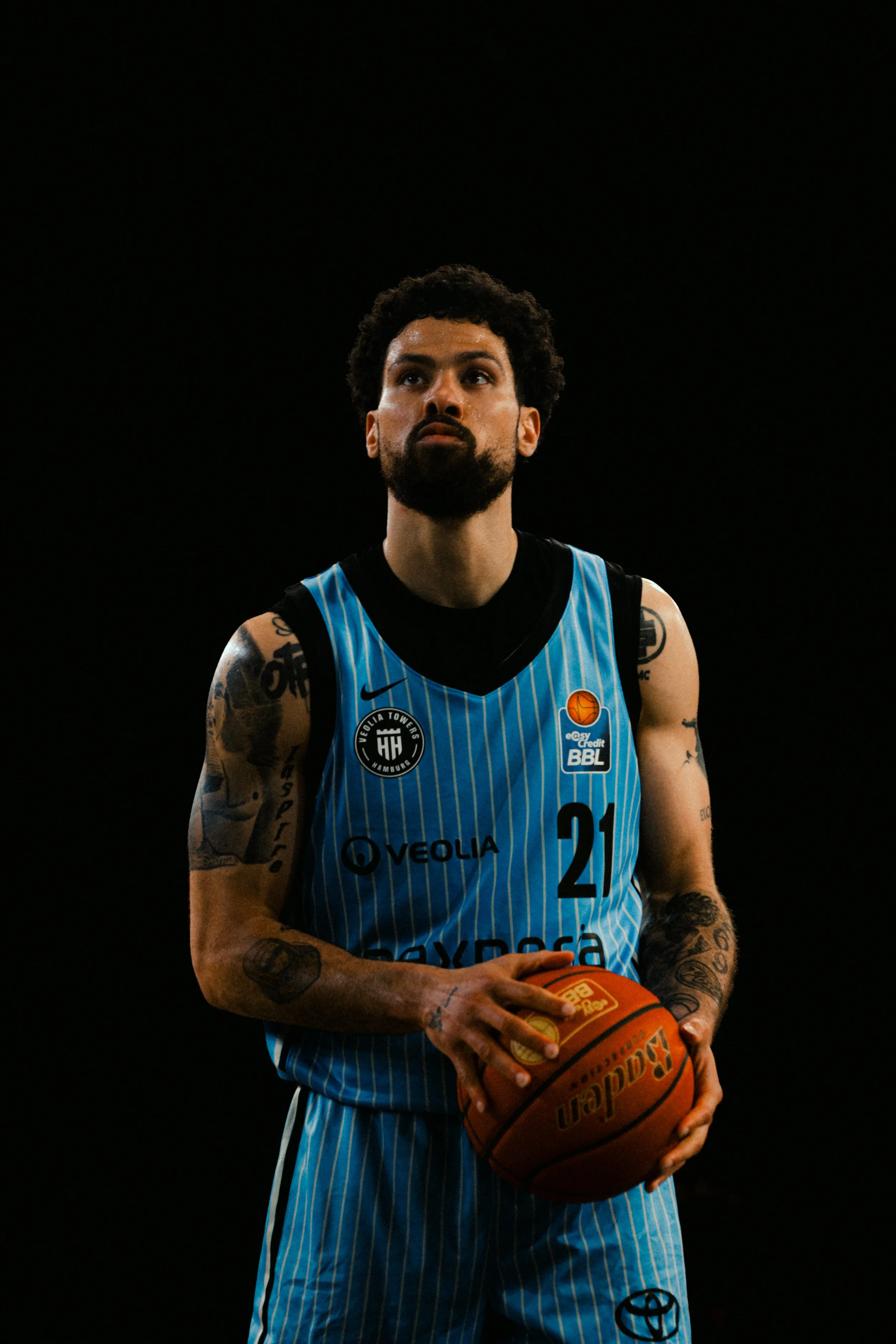 A male basketball player with tattoos on his arms, wearing a blue striped jersey, holding a basketball, standing against a black background.