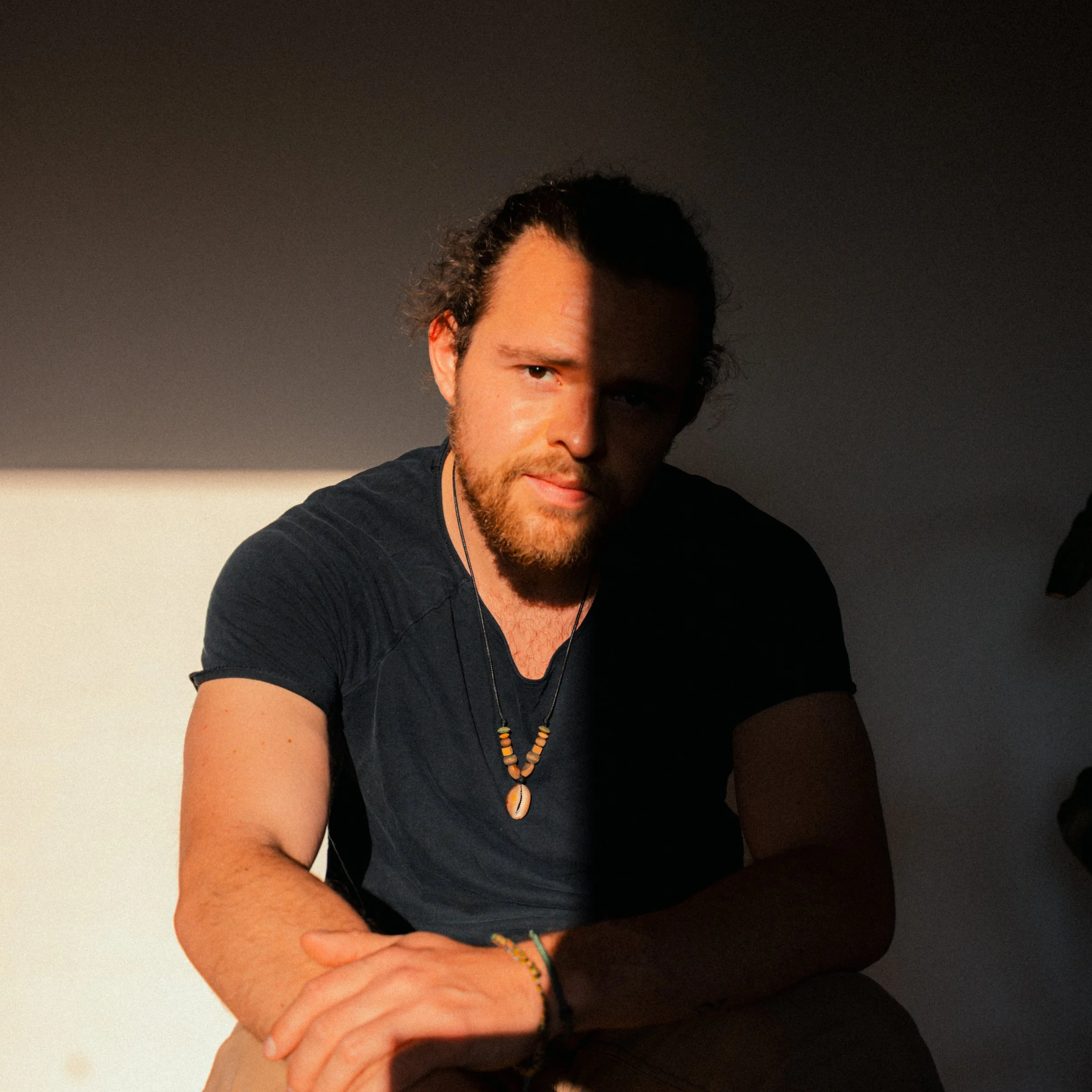 A young man with long, curly hair and a beard, wearing a black shirt and beaded necklaces, sitting with his arms crossed in front of a background that is dark on top and light on the bottom.