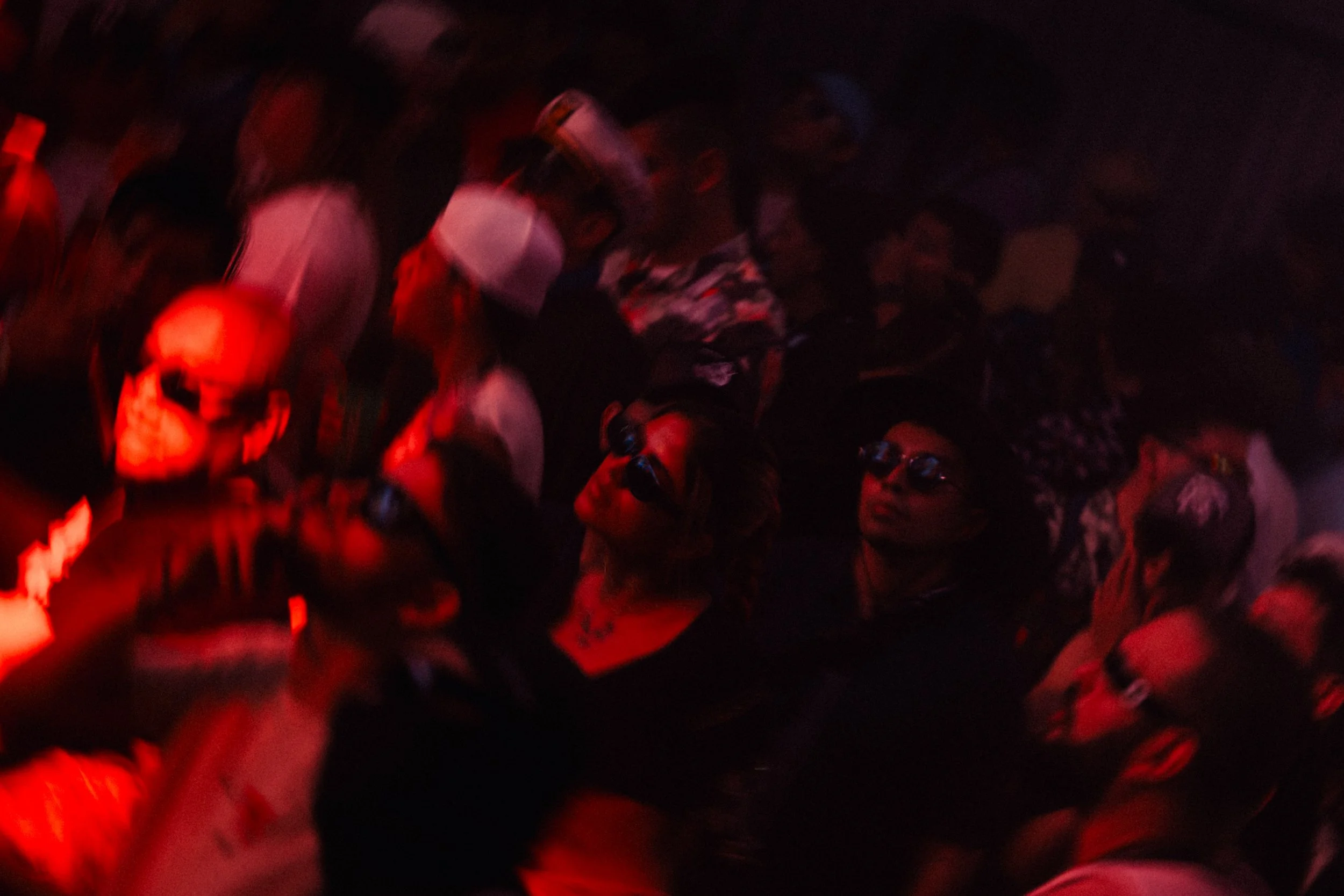 A dark scene with people gathered closely together, illuminated by red light, wearing glasses and casual clothing, with some faces partially visible.