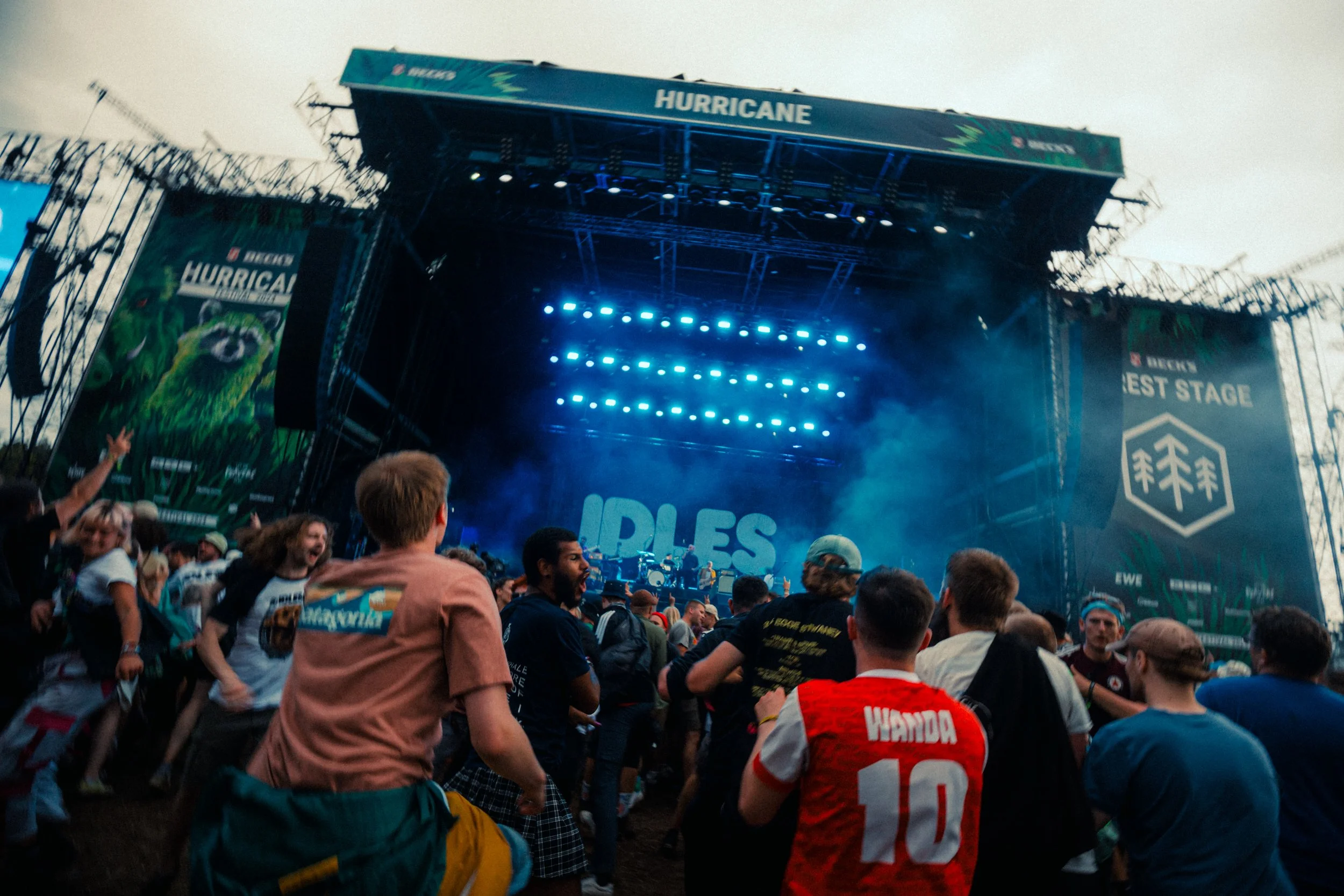 Crowd of people at an outdoor concert or music festival with a large stage labeled 'HURRICANE' and banners, some wearing jerseys, some dancing and enjoying the event.
