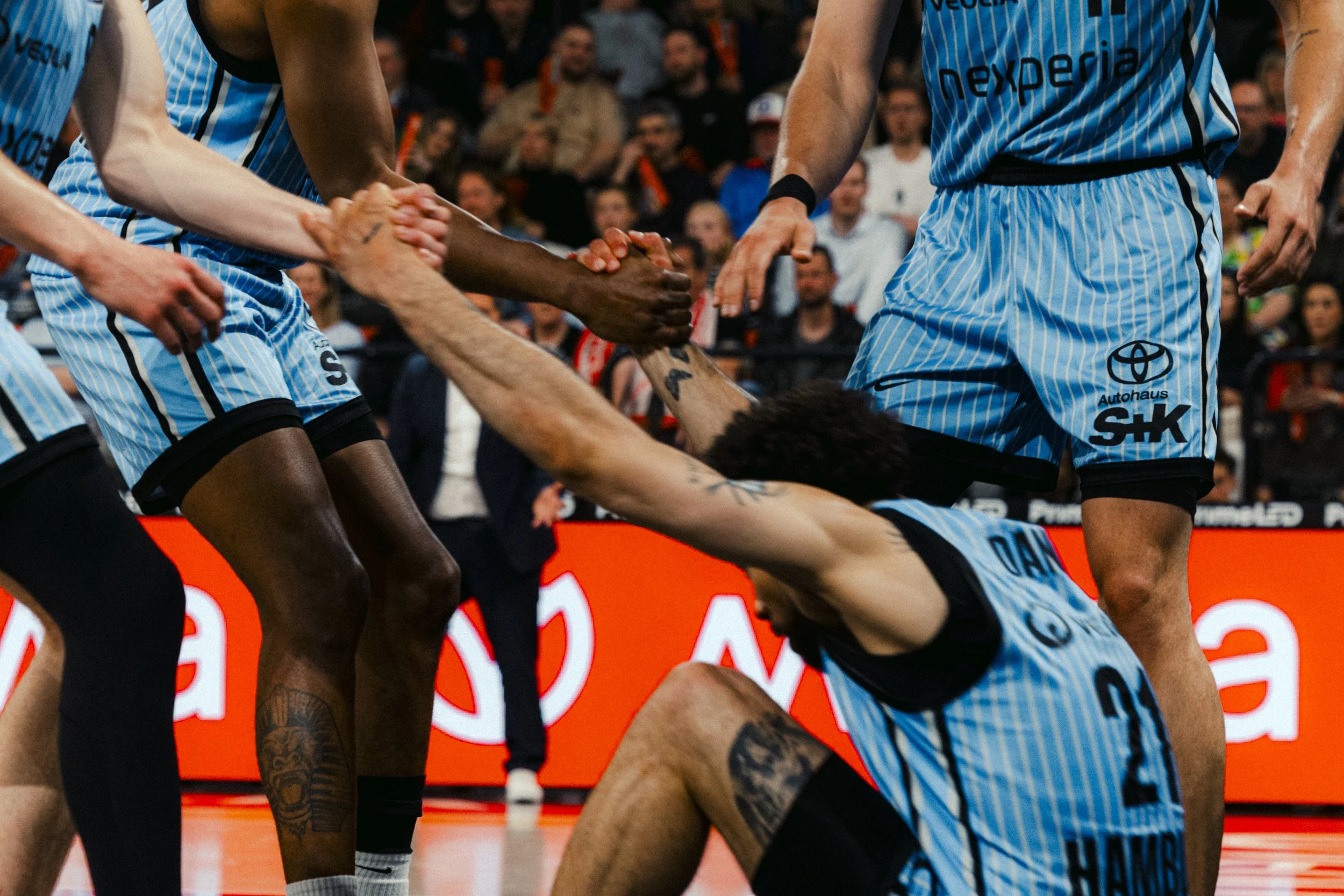 Basketball players in blue uniforms are participating in a game, with some players celebrating or in action on the court, and spectators watching in the background.