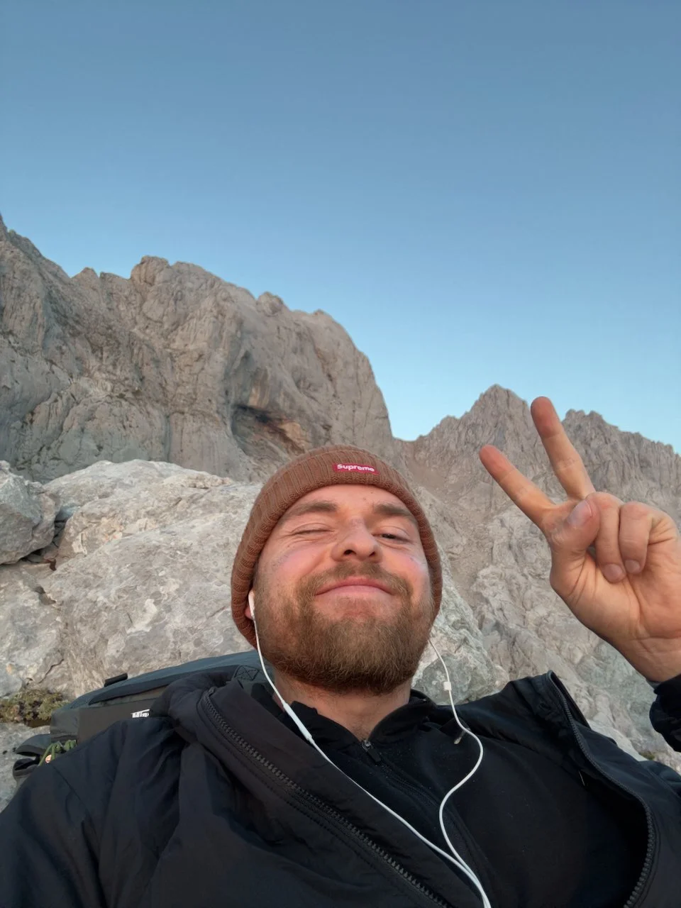 A man taking a selfie outdoors against a mountain backdrop, wearing a brown beanie, black jacket, earphones, and smiling while flashing a peace sign.