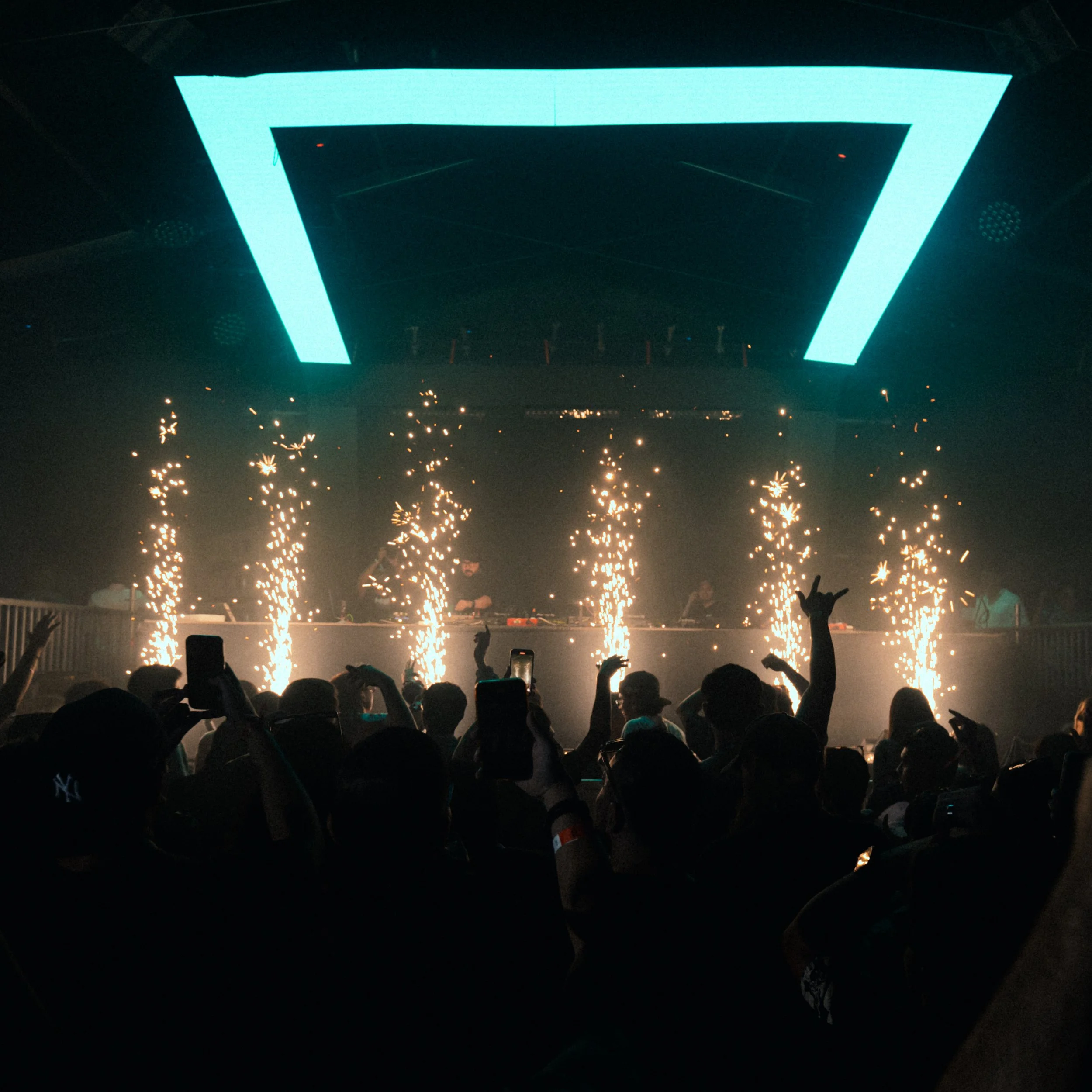 Nightclub scene with a large illuminated geometric display hanging from the ceiling, sparklers in the foreground, and a crowd of people with raised hands and smartphones capturing the moment.
