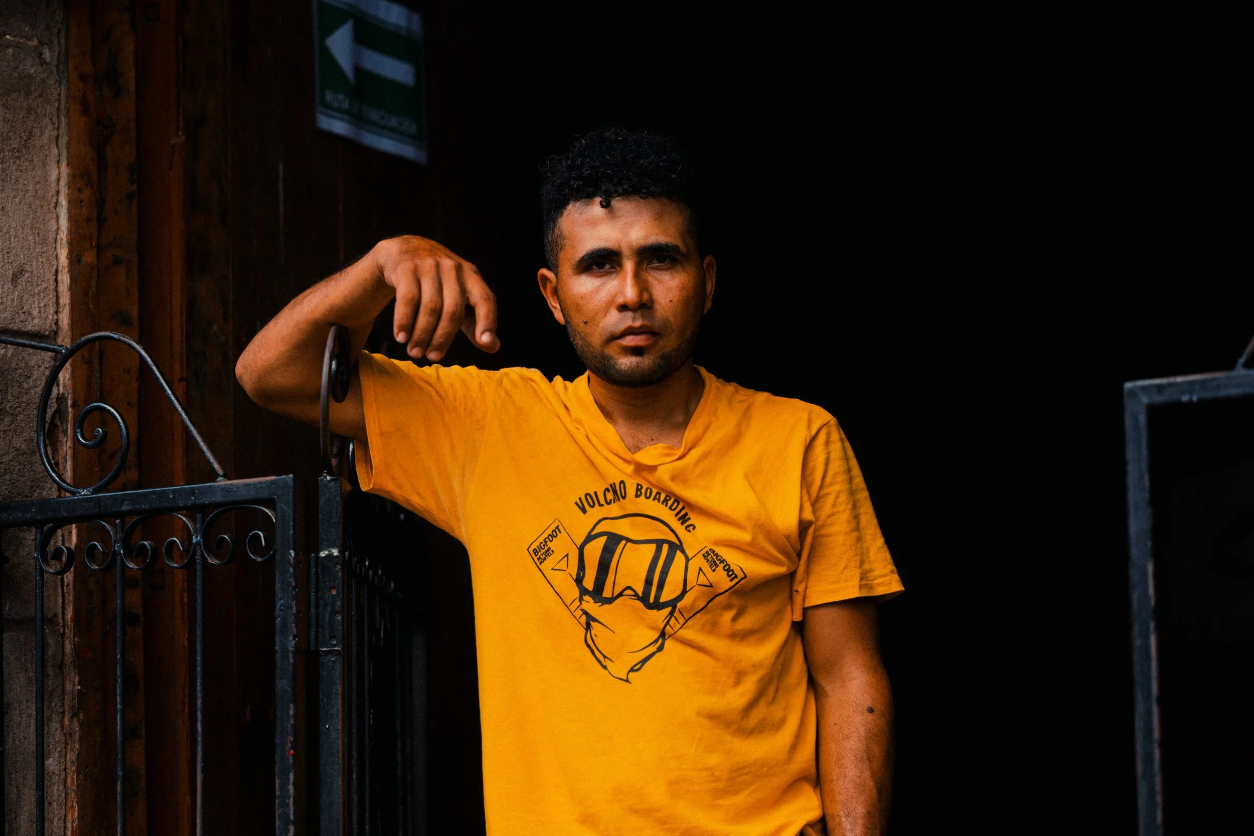 A man with curly black hair and a beard, wearing a yellow t-shirt with a graphic of a face and the words 'Volcom Boarding' and 'Bigfoot', leaning on a black metal fence with a serious expression against a dark background.