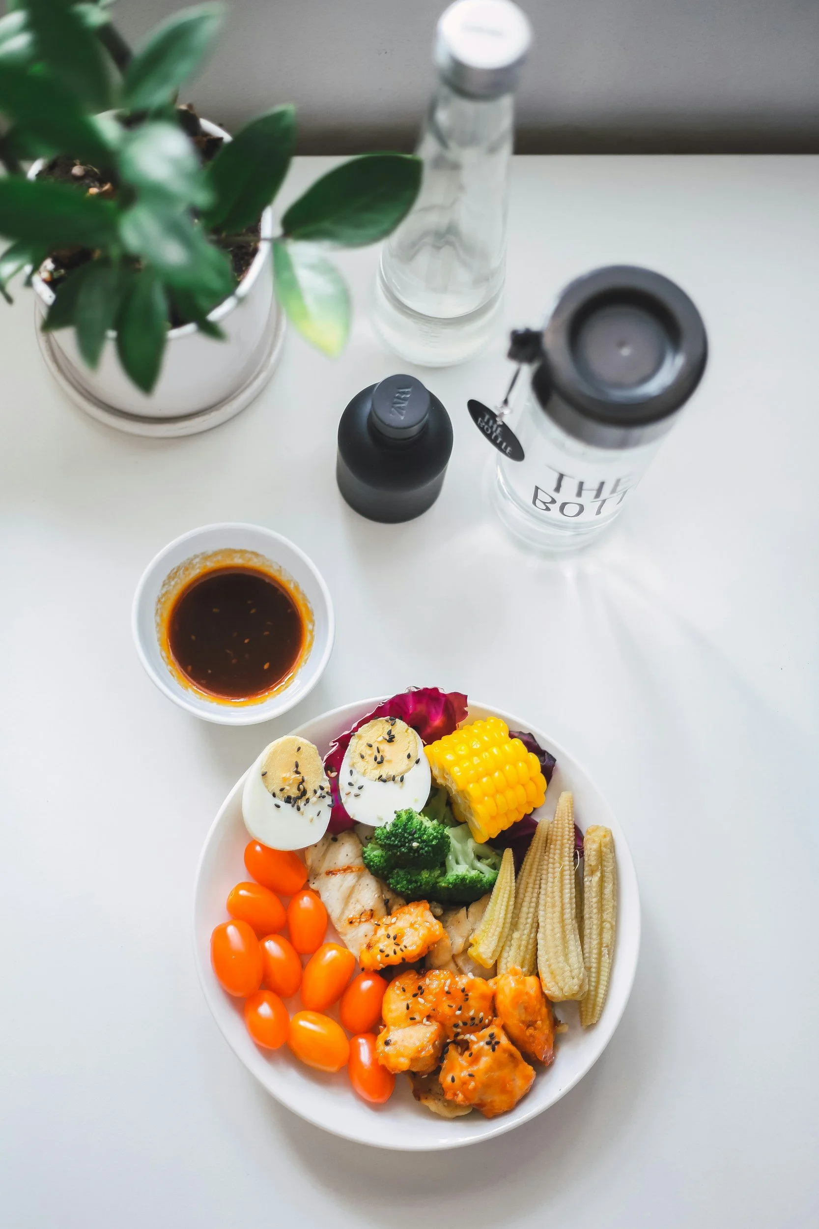 Healthy vegetable platter with grilled chicken, eggs, cherry tomatoes, broccoli, corn, baby corn, dipping sauce, and water bottle on a white table
