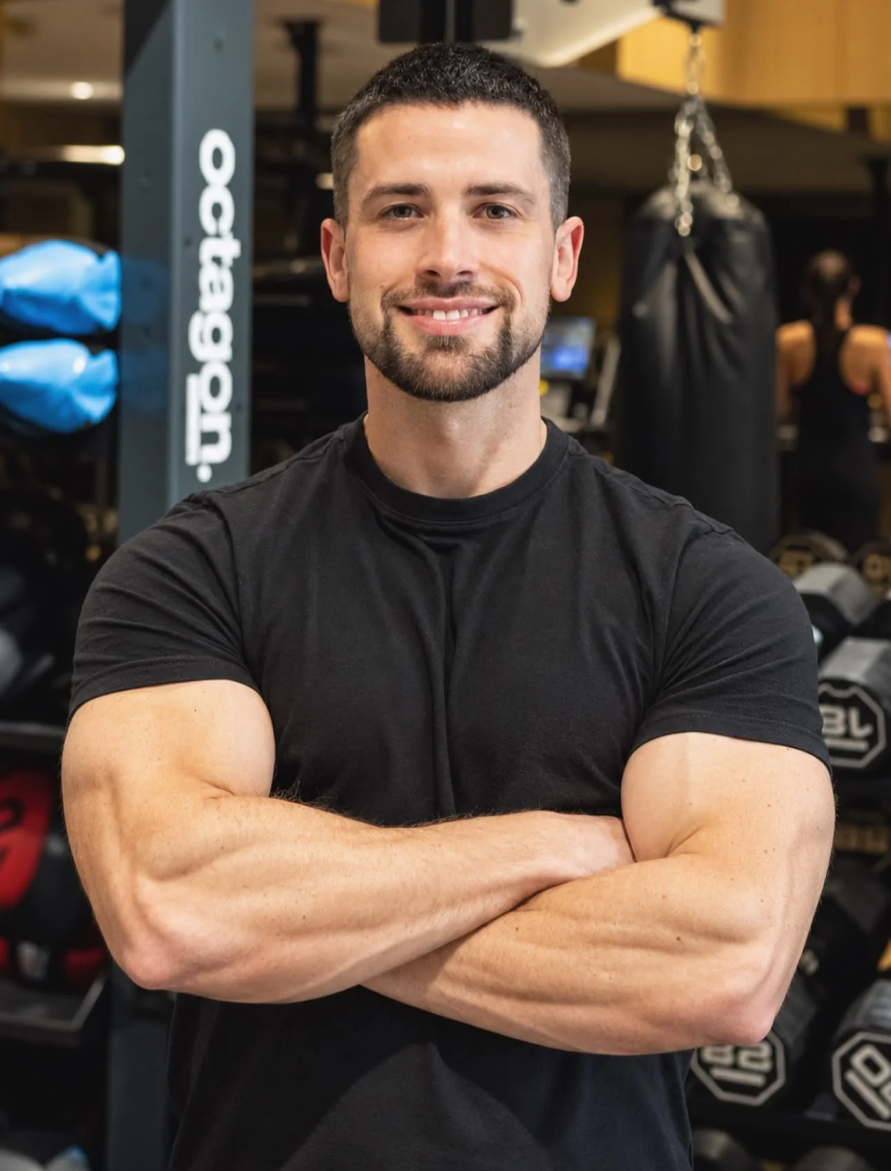 Muscular man in a black T-shirt standing with crossed arms in a gym, with punching bags and weights in the background.